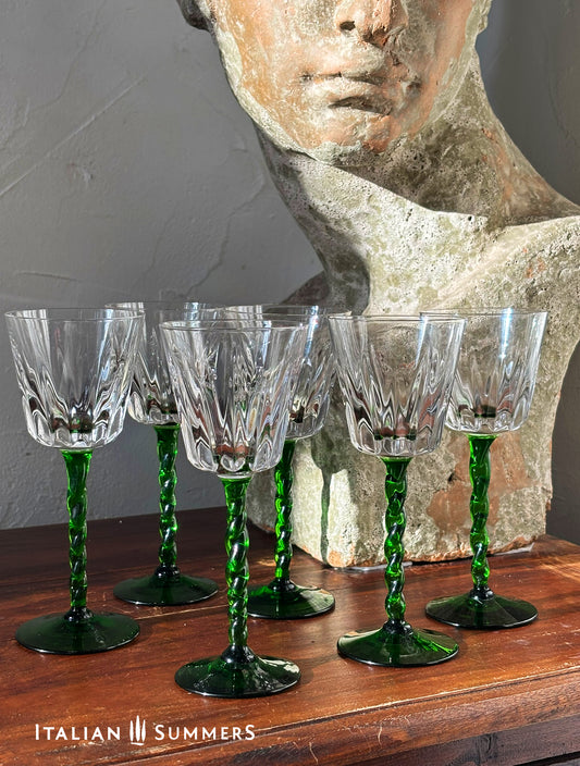 Set of six crystal wine glasses with green stems on a wooden surface, with a stone sculpture in the background.