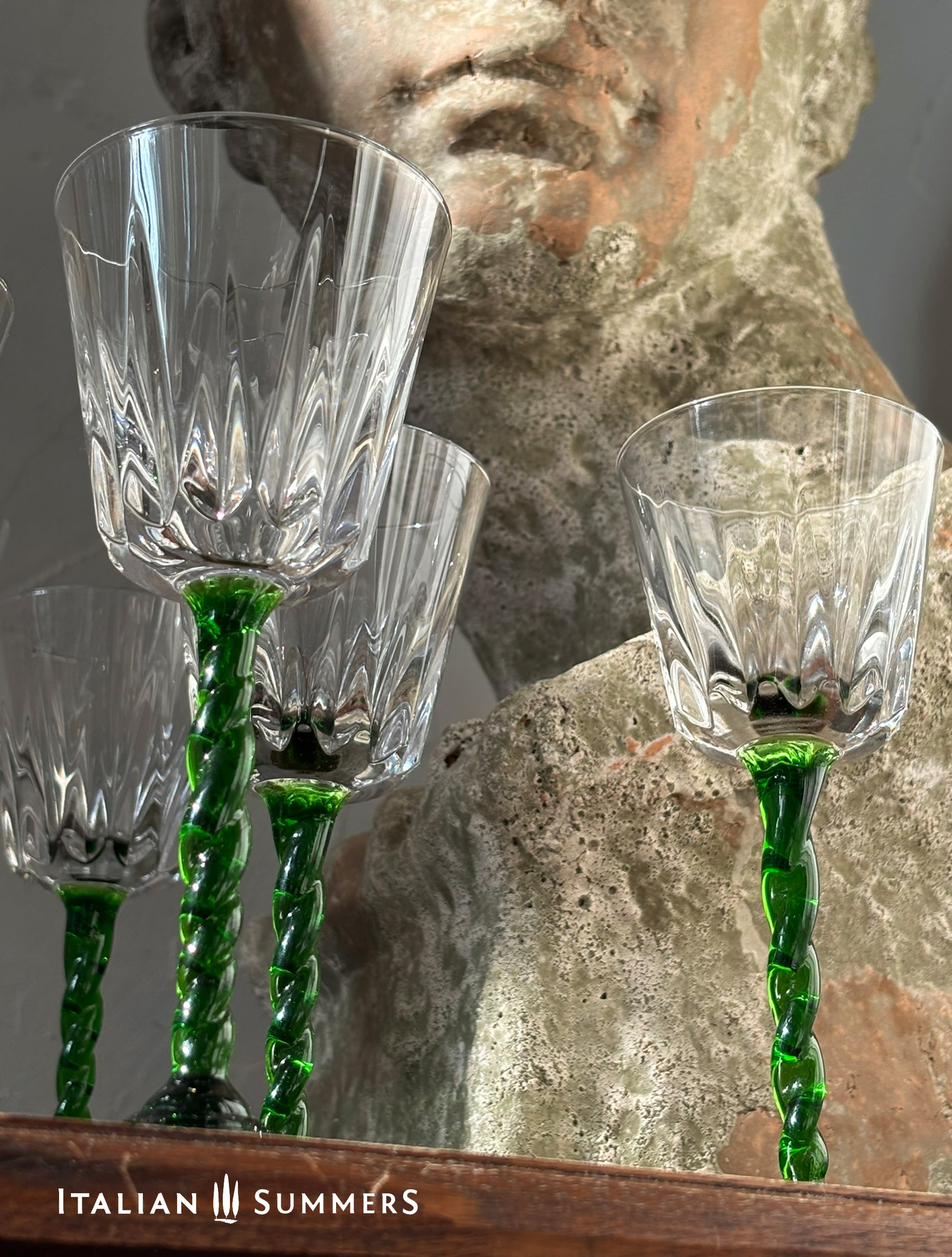 Set of crystal wine glasses with green stems in front of a stone sculpture.