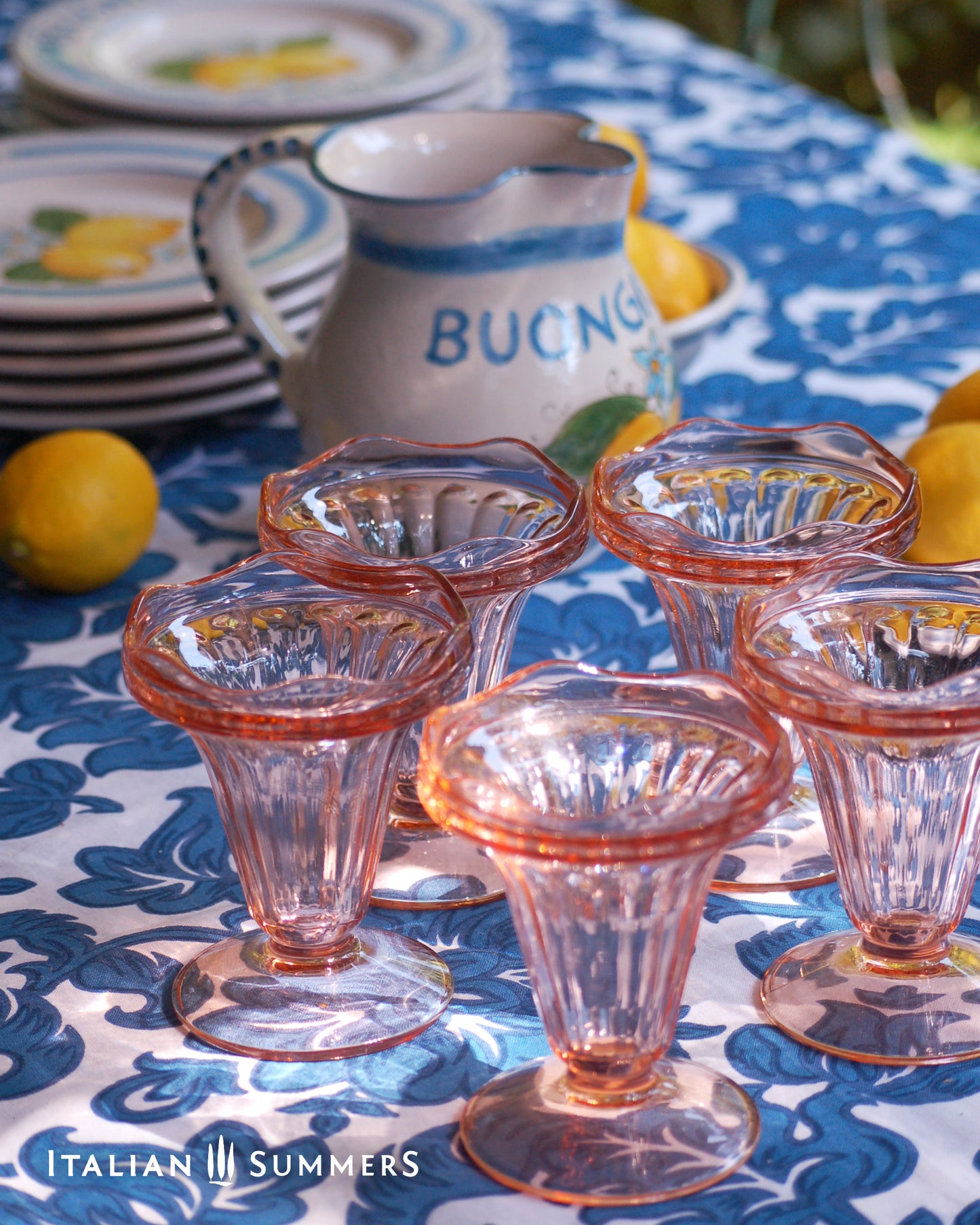 Set of vintage French pink glass dessert coupes  on a blue and white floral patterned tablecloth  with lemons and a white ad blue ceramic carafe