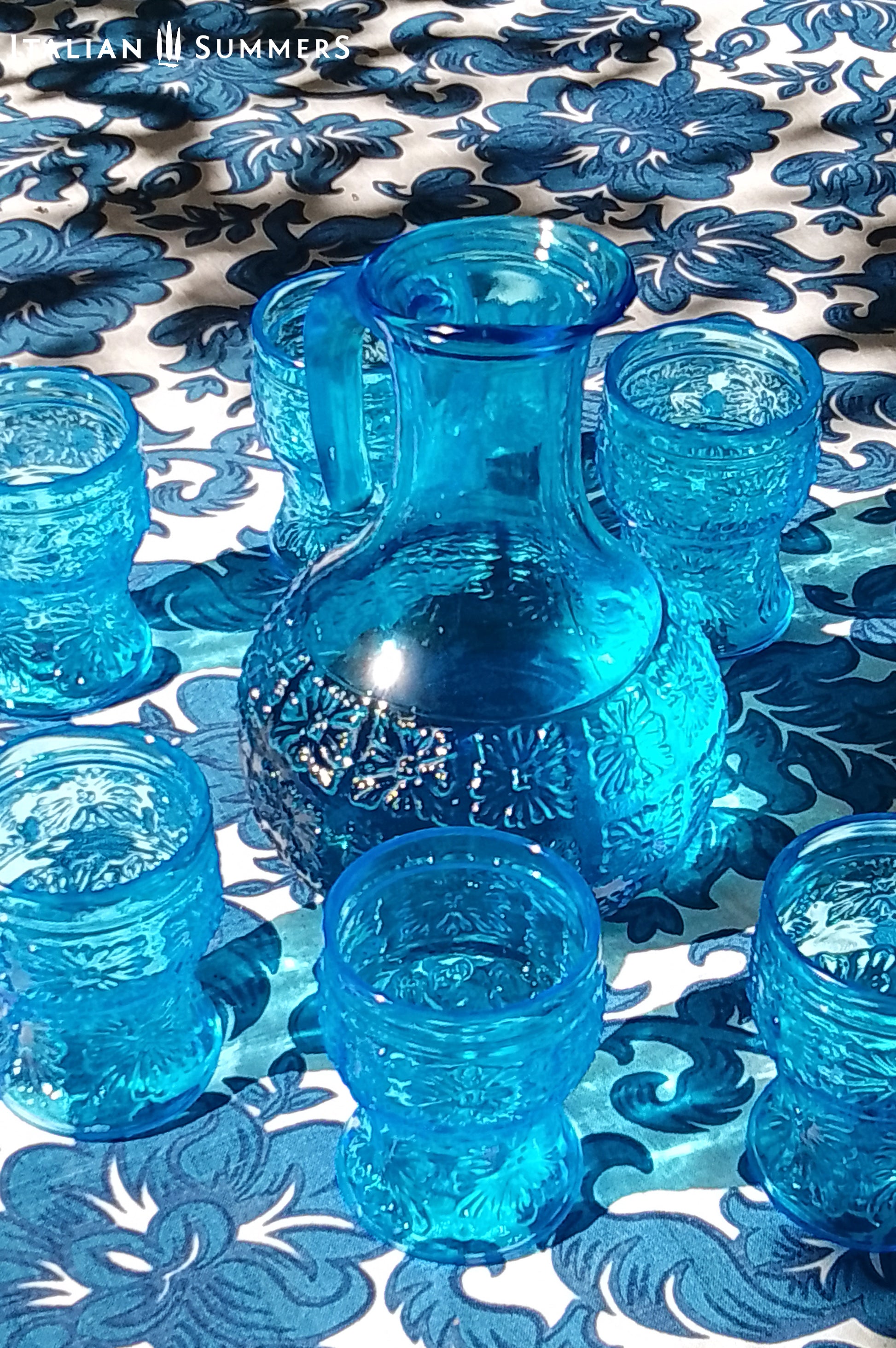 A blue glass carafe and six tumblers set, all embossed in flower patterns from the 1970s Italy, sits on a table with a colorful and bright tablecloth with blue floral patterns.