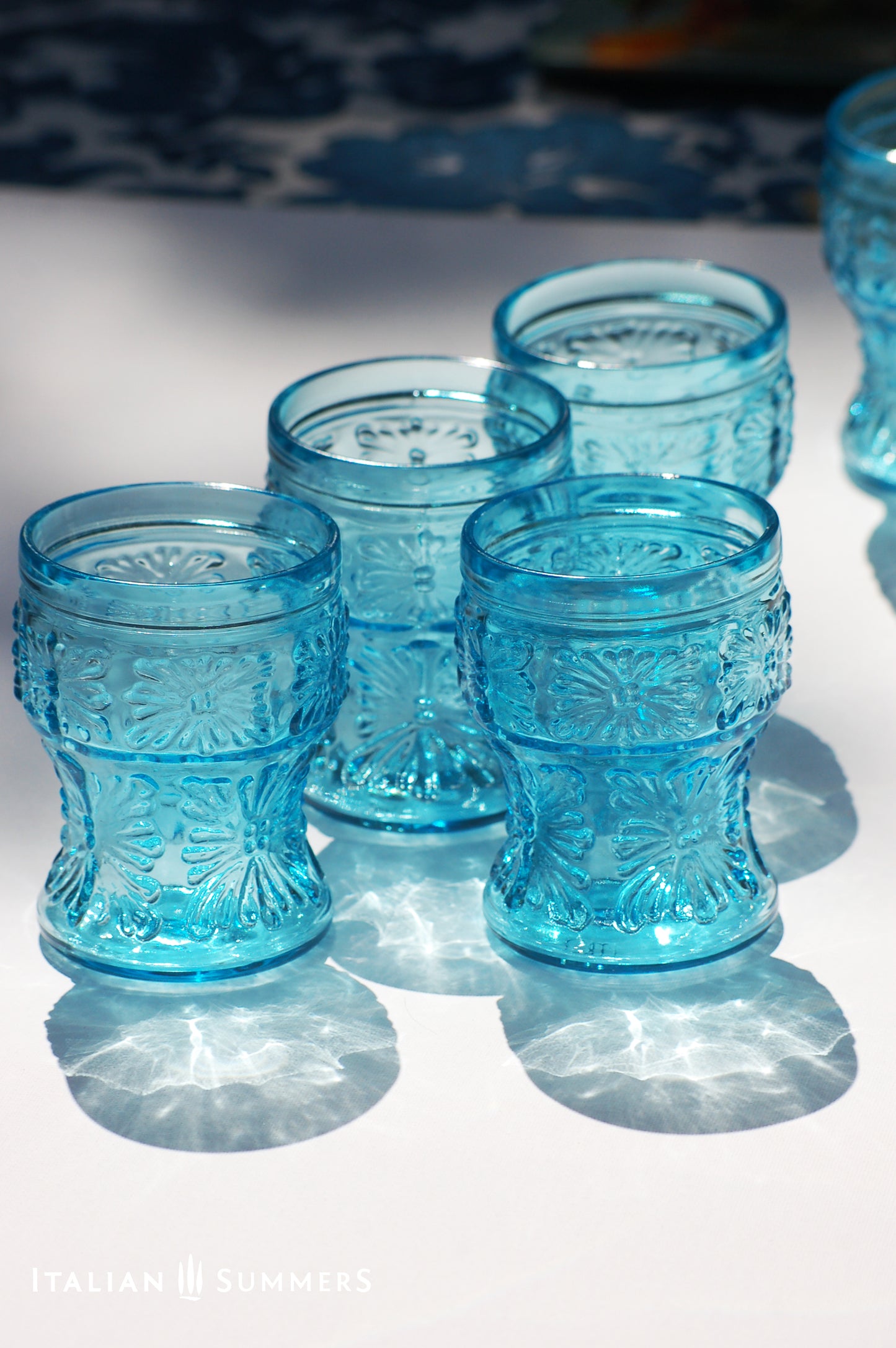 A blue glass carafe and six tumblers set, all embossed in flower patterns from the 1970s Italy, sits on a table with a colorful and bright tablecloth with blue floral patterns.