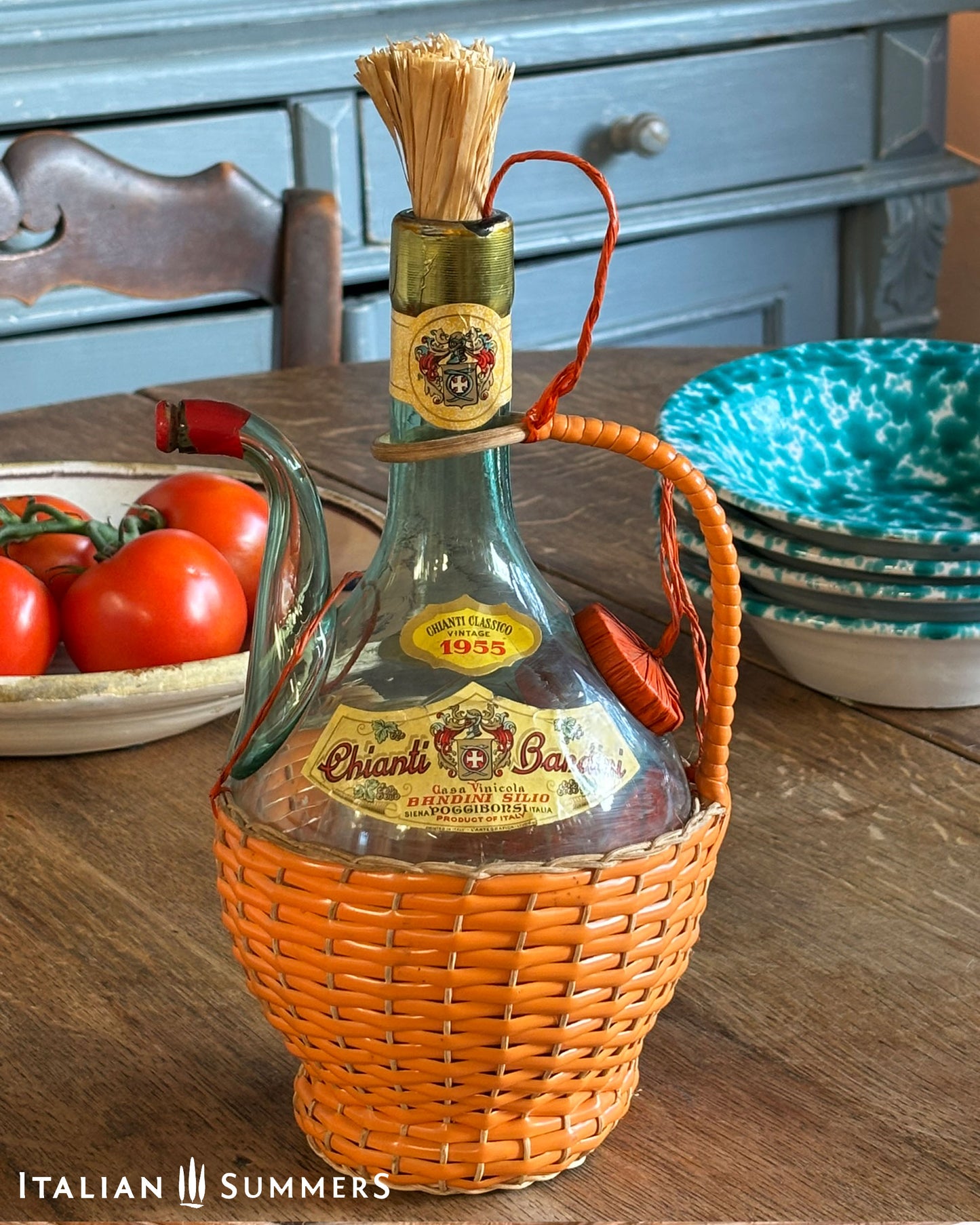 Vintage 1955 bottle of Chianti Wine in an orange basket on a wooden table with tomatoes and plates in the background.