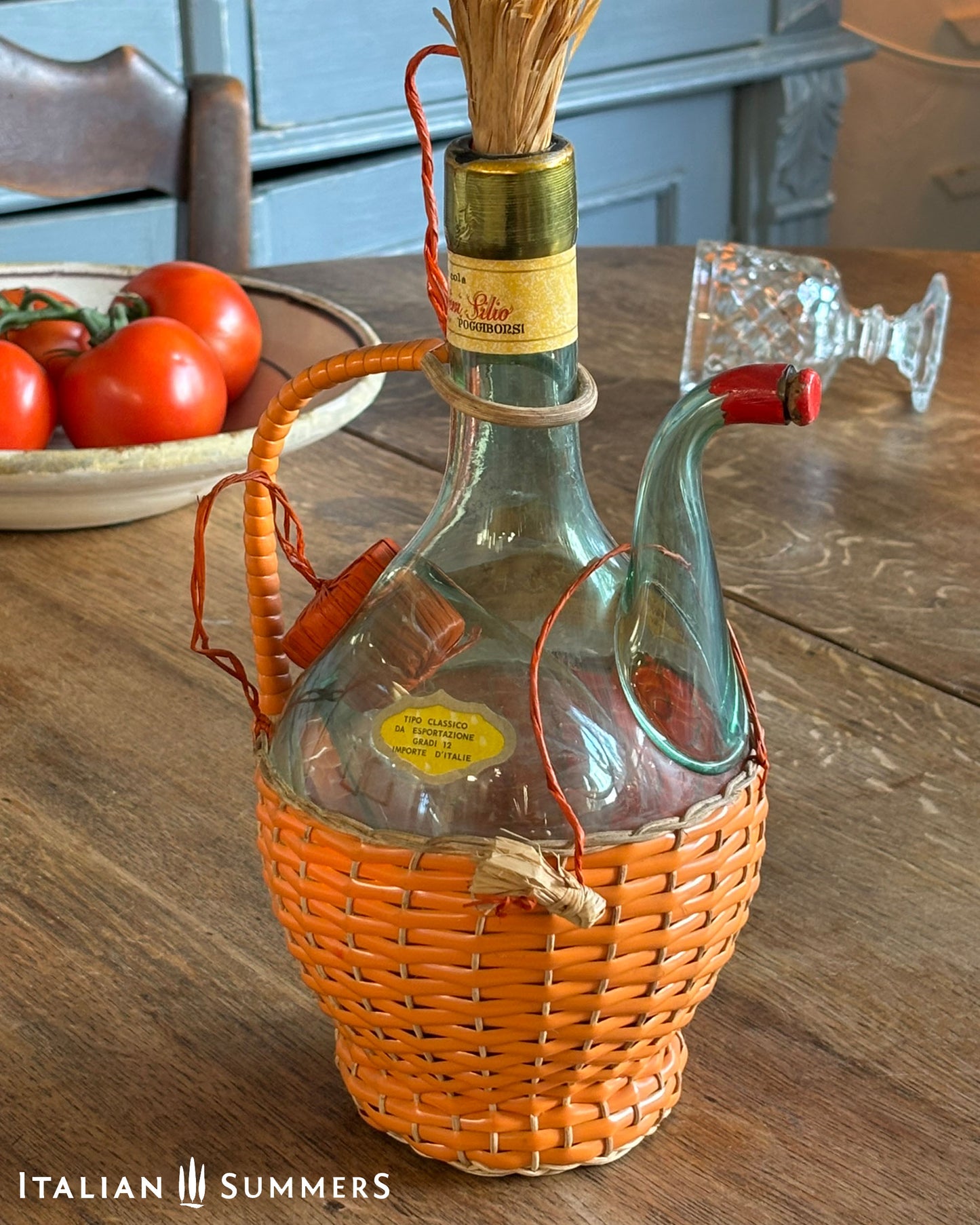 Decorative bottle with wicker basket on a wooden table, tomatoes in the background.