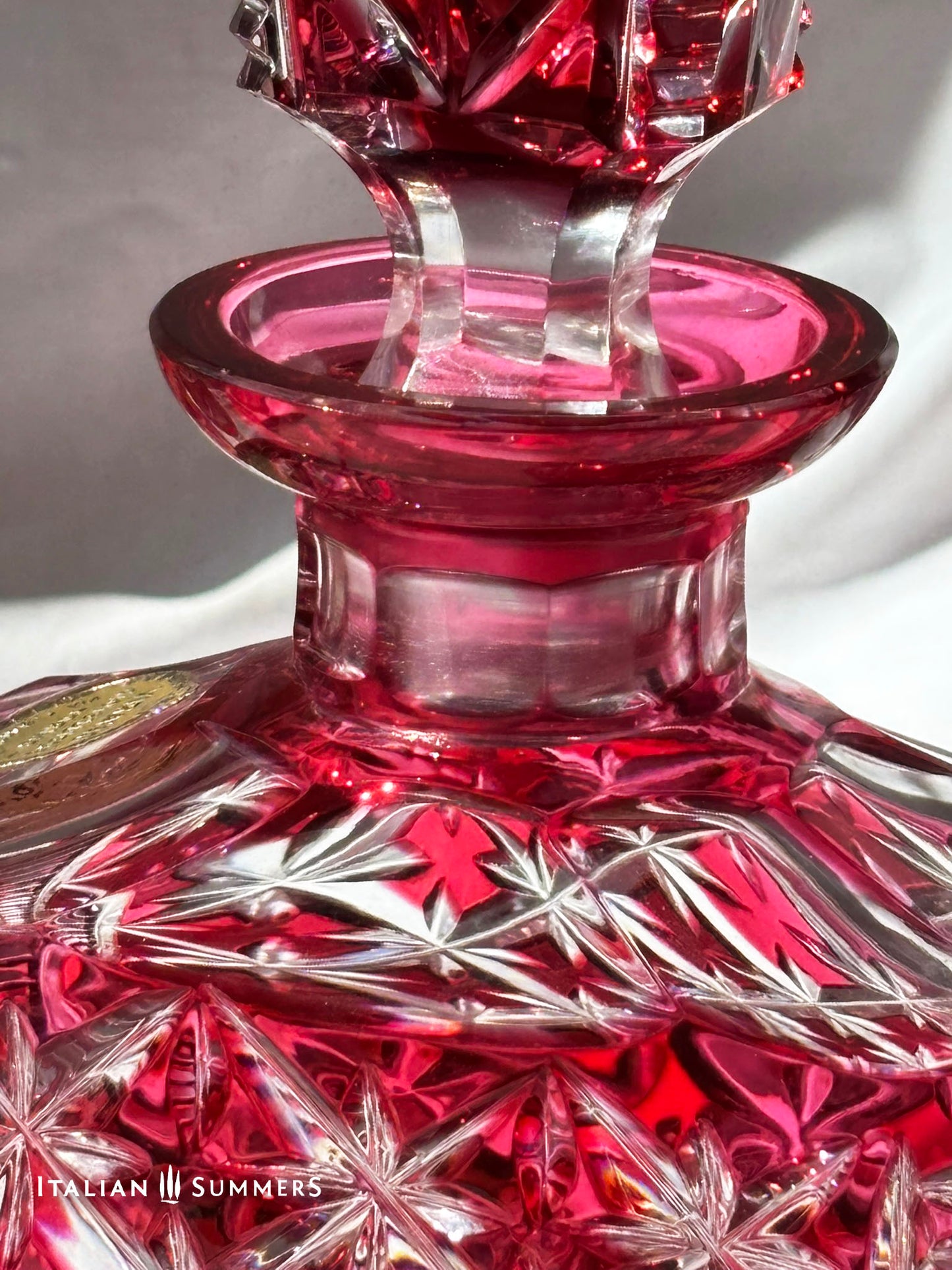 deail of a stopper from a cut crystal cranberry-red decanter The glassware is displayed on a white surface. The text "ITALIAN SUMMERS" is visible.