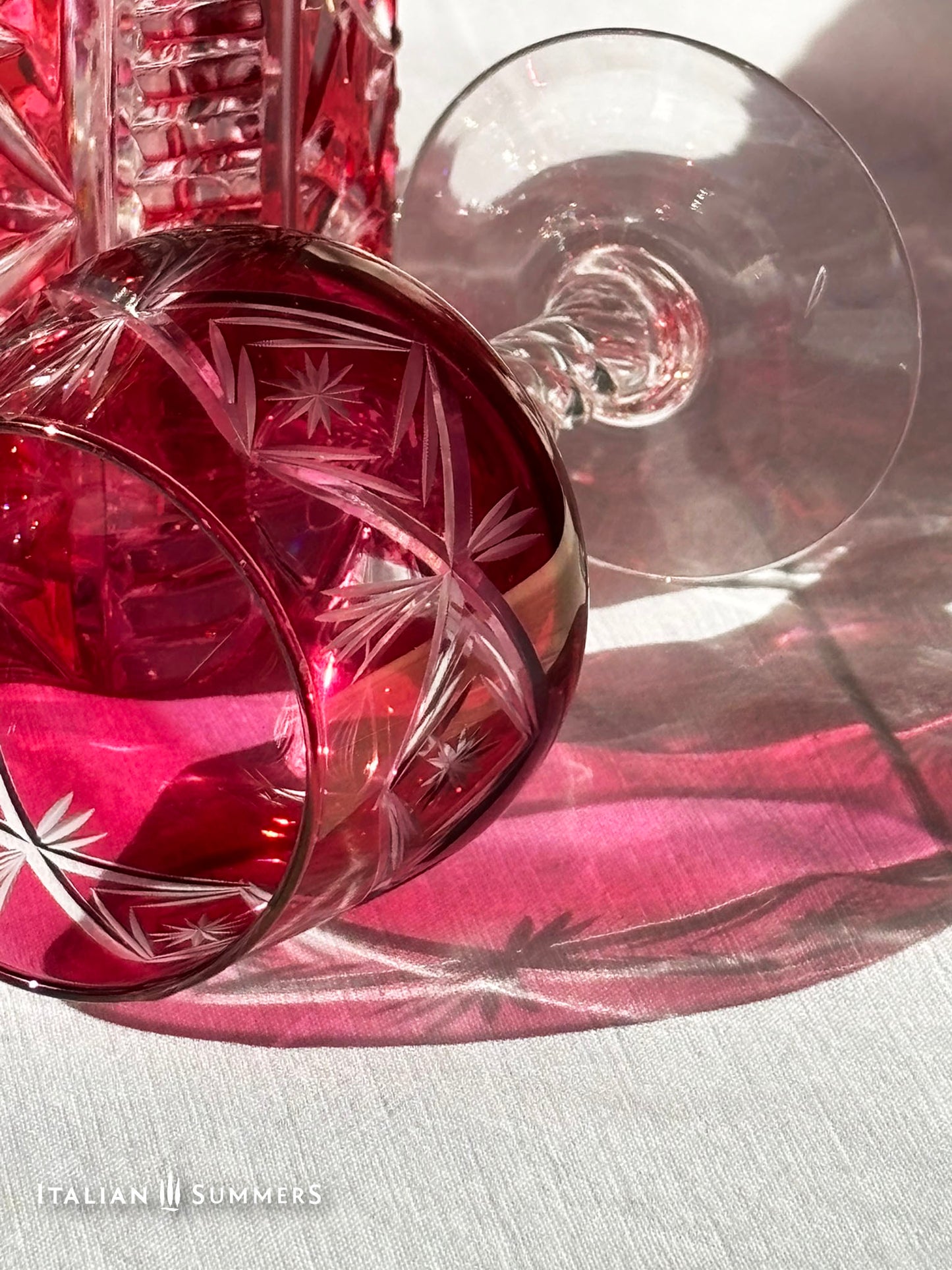 A set of cranberry red crystal glassware featuring a geometric starburst design. The set includes a rectangular decanter with a stopper and several wine glasses. The glassware is displayed on a white surface. The text "ITALIAN SUMMERS" is visible.