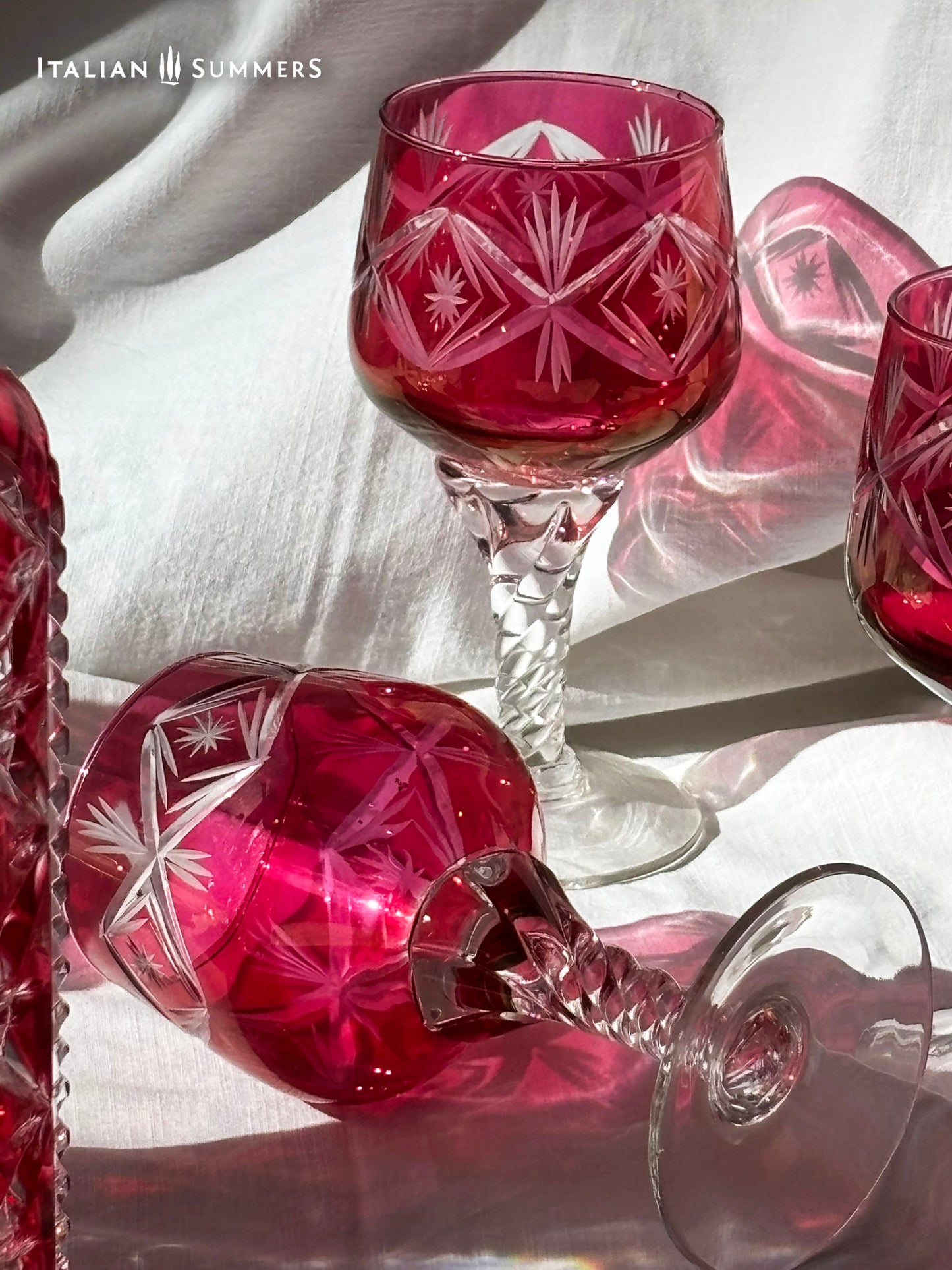  A set of cranberry red crystal glassware featuring a geometric starburst design. The set includes a rectangular decanter with a stopper and several wine glasses. The glassware is displayed on a white surface. The text "ITALIAN SUMMERS" is visible.