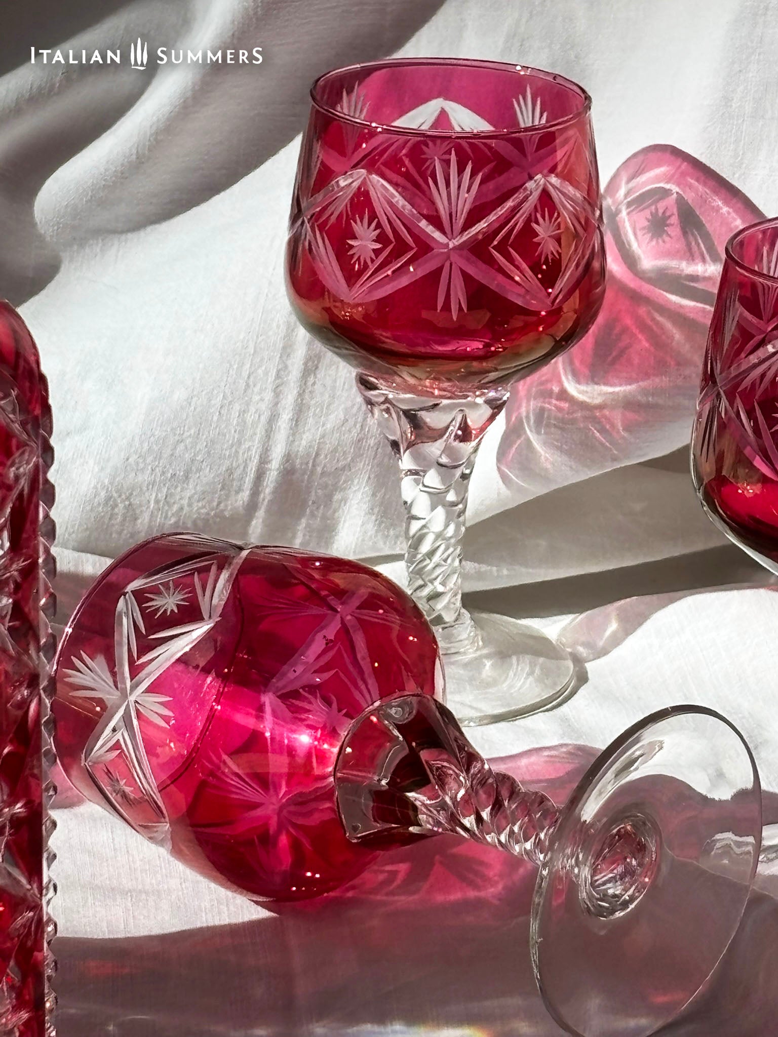  A set of cranberry red crystal glassware featuring a geometric starburst design. The set includes a rectangular decanter with a stopper and several wine glasses. The glassware is displayed on a white surface. The text "ITALIAN SUMMERS" is visible.
