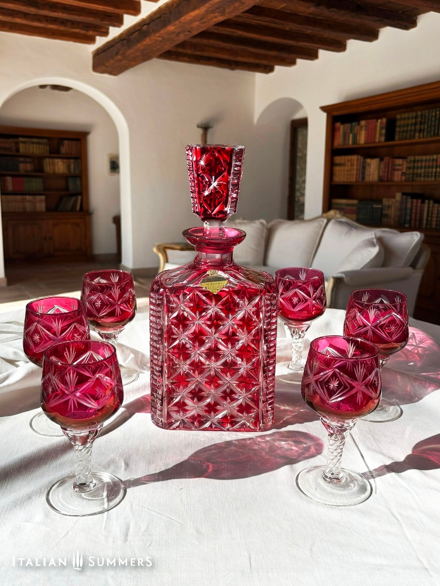  A set of cranberry red crystal glassware featuring a geometric starburst design. The set includes a rectangular decanter with a stopper and several wine glasses. The glassware is displayed on a white surface. The text "ITALIAN SUMMERS" is visible.