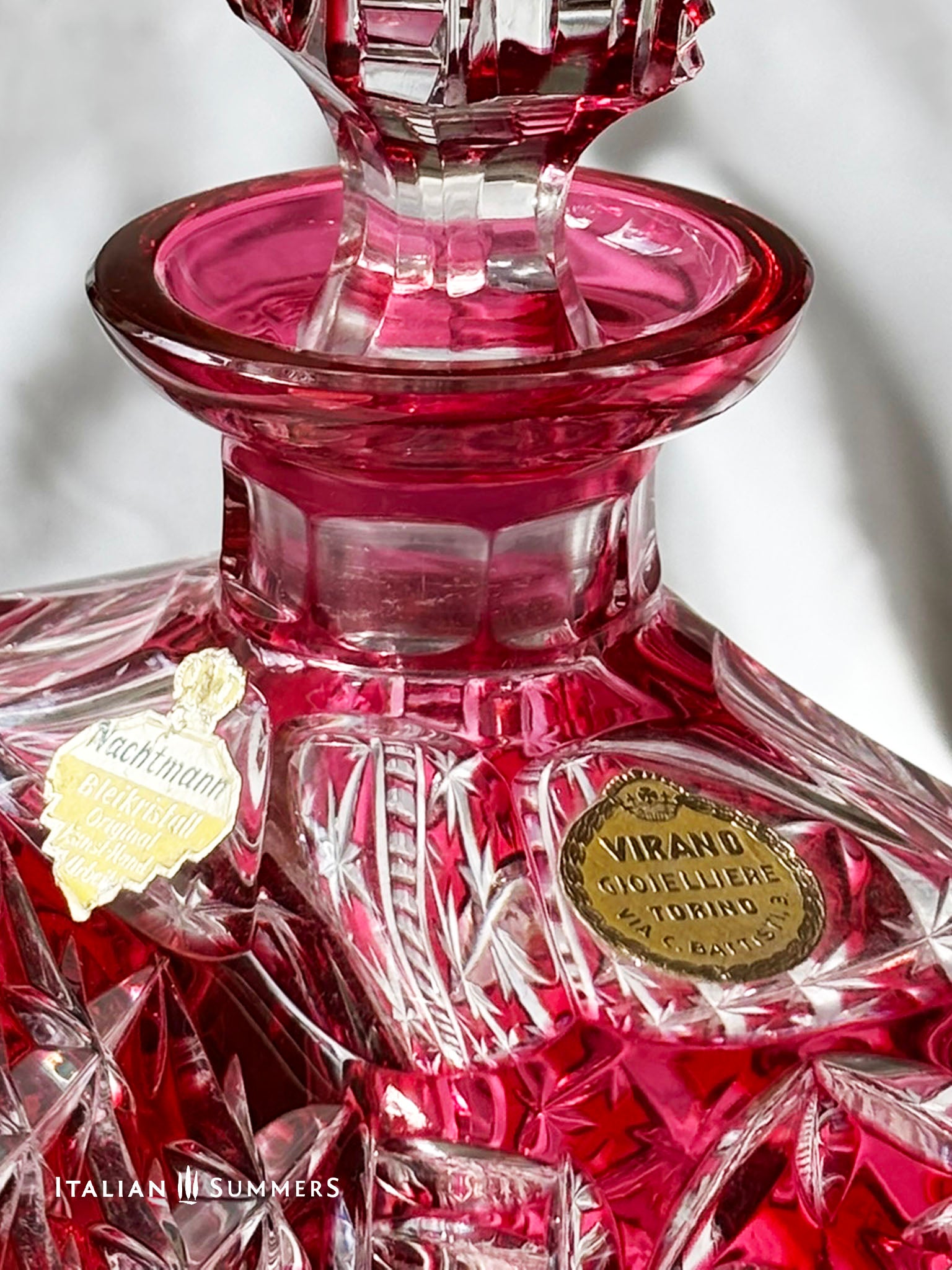  A set of cranberry red crystal glassware featuring a geometric starburst design. The set includes a rectangular decanter with a stopper and several wine glasses. The glassware is displayed on a white surface. The text "ITALIAN SUMMERS" is visible.