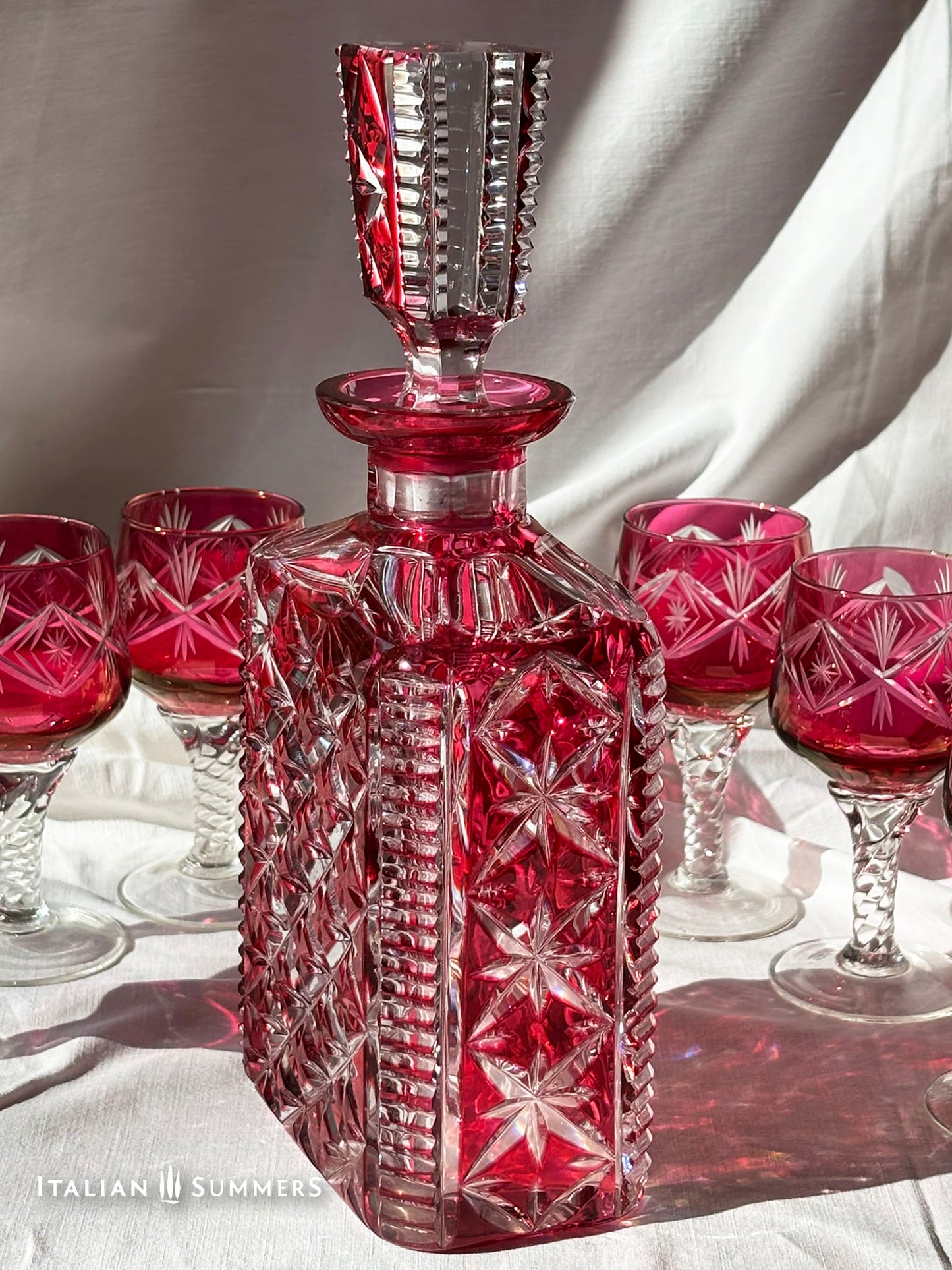 A set of ruby red crystal glassware, including a decanter with a stopper and six wine glasses. The decanter and glasses have a diamond pattern. The glasses have clear, twisted stems. The set is on a white surface.