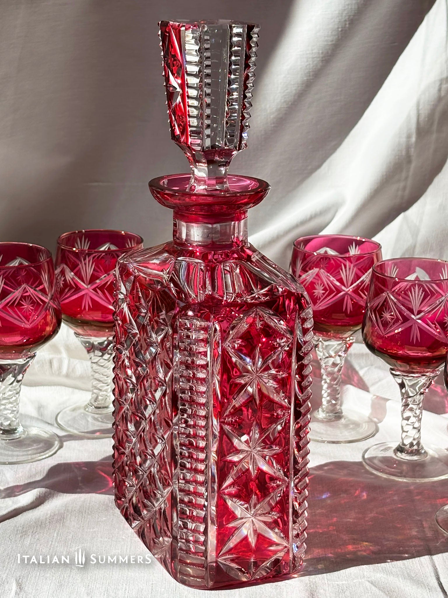 A set of ruby red crystal glassware, including a decanter with a stopper and six wine glasses. The decanter and glasses have a diamond pattern. The glasses have clear, twisted stems. The set is on a white surface.