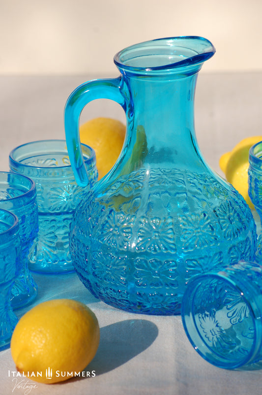 A blue glass carafe and six tumblers set, all embossed in flower patterns from the 1970s Italy, sits on a table with a colorful and bright tablecloth with blue floral patterns.