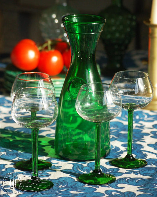 An emerald green glass carafe stands surrounded by four green-stemmed wine  glasses on  top of  a blue and white floral tablecloth with colorful ceramic plates with bright tomatoes in the background