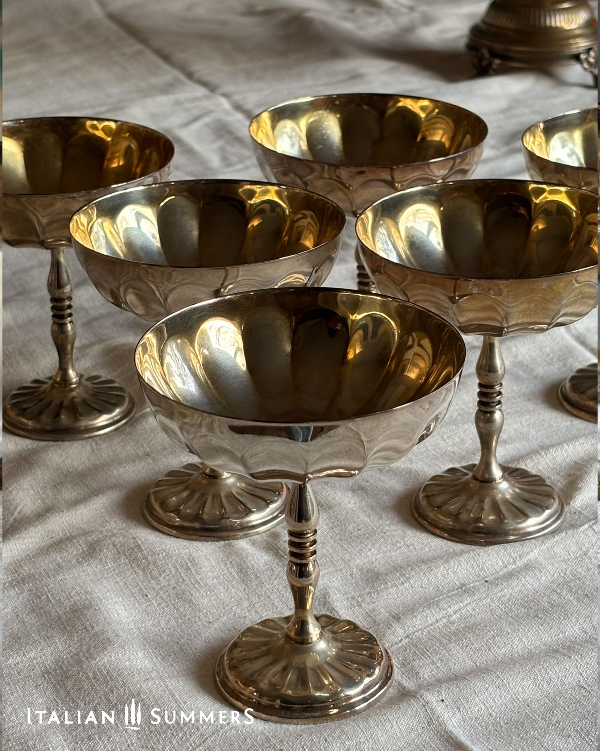Set of silver stemmed champagne coupes on a textured surface with 'Italian Summers' branding.