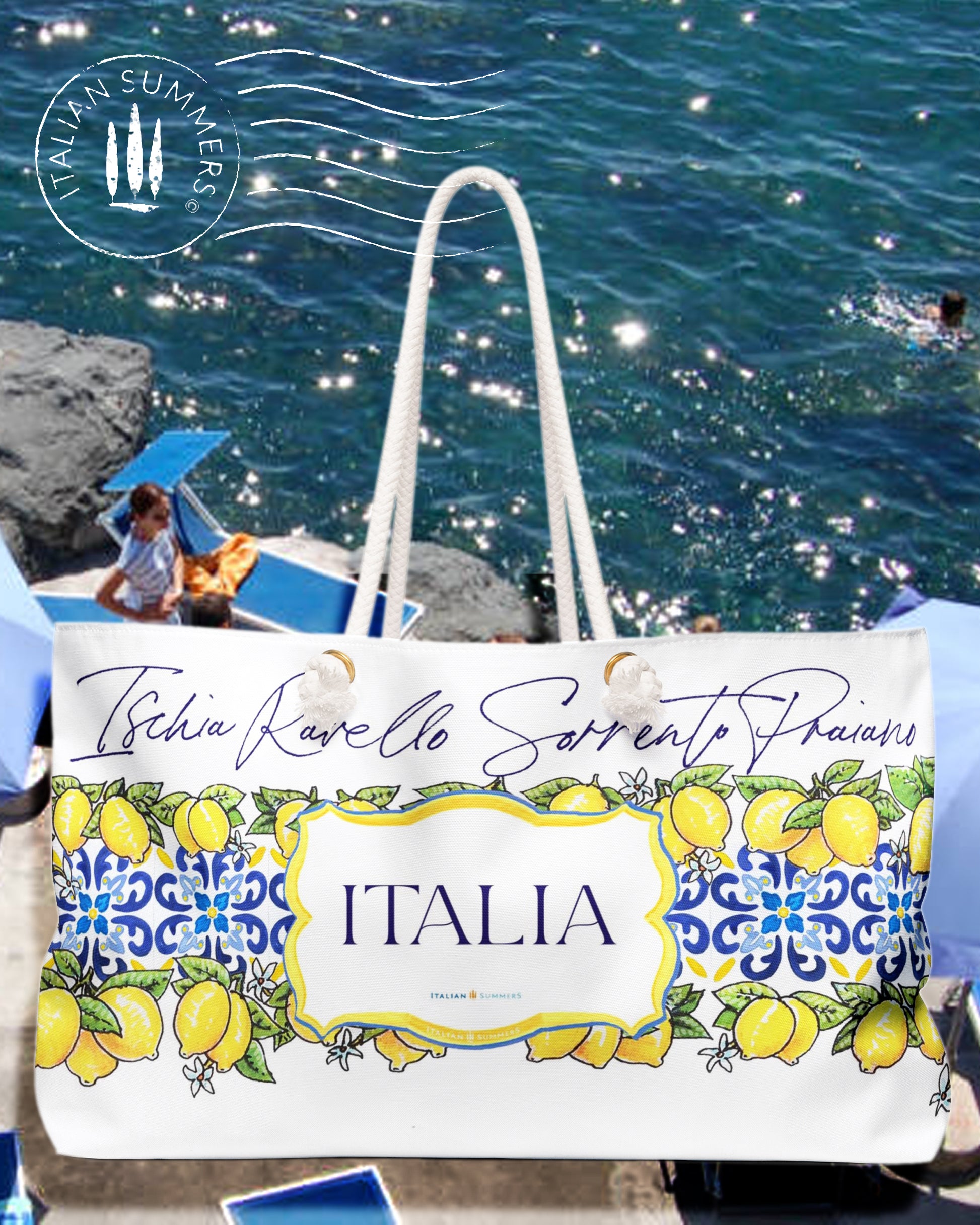 Italy-inspired XL Beach Bag  with printed blue Italian tiles in a  large band across the entire bag , framed with garlands of sunny, leafy Sorrento Lemons  with flowers. A large cartouche -like crest in the middle has the word  ITALIA - Seaside location names are writthen in flowing hand-script in blue across the top of the design. Sorrento, Positano, Ravello, Praiano, Capri, Amalfi, Procida,  made by Italian Summers