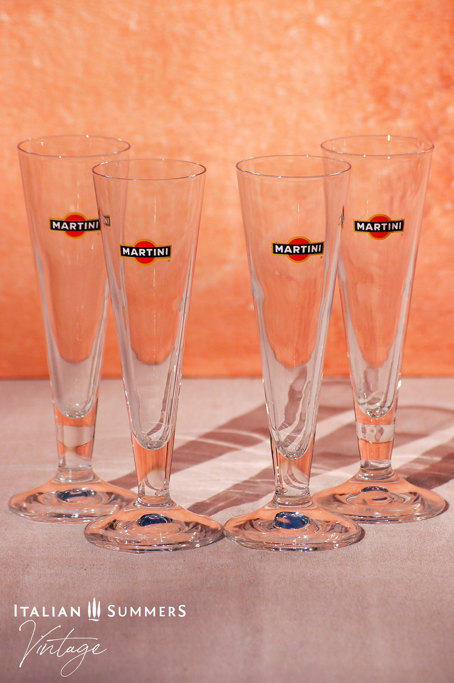Set of four elegant vintage Martini glasses, each featuring the iconic Martini logo on the front. The glasses are displayed on a book titled 'Dolce Vita Style', set against a warm, peach-colored background with the text 'Italian Summers Vintage'.