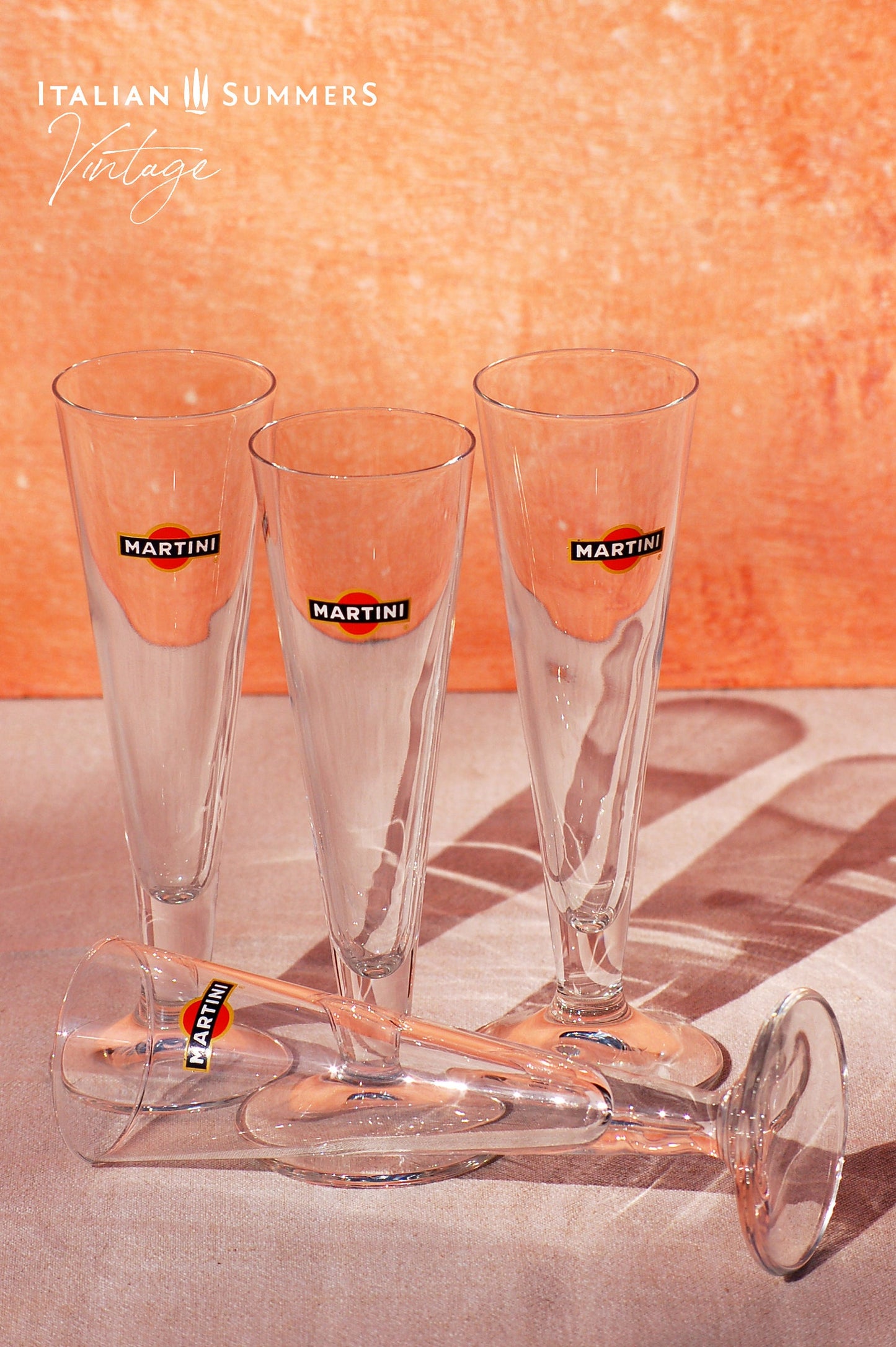 Set of four elegant vintage Martini glasses, each featuring the iconic Martini logo on the front. The glasses are displayed on a book titled 'Dolce Vita Style', set against a warm, peach-colored background with the text 'Italian Summers Vintage'.