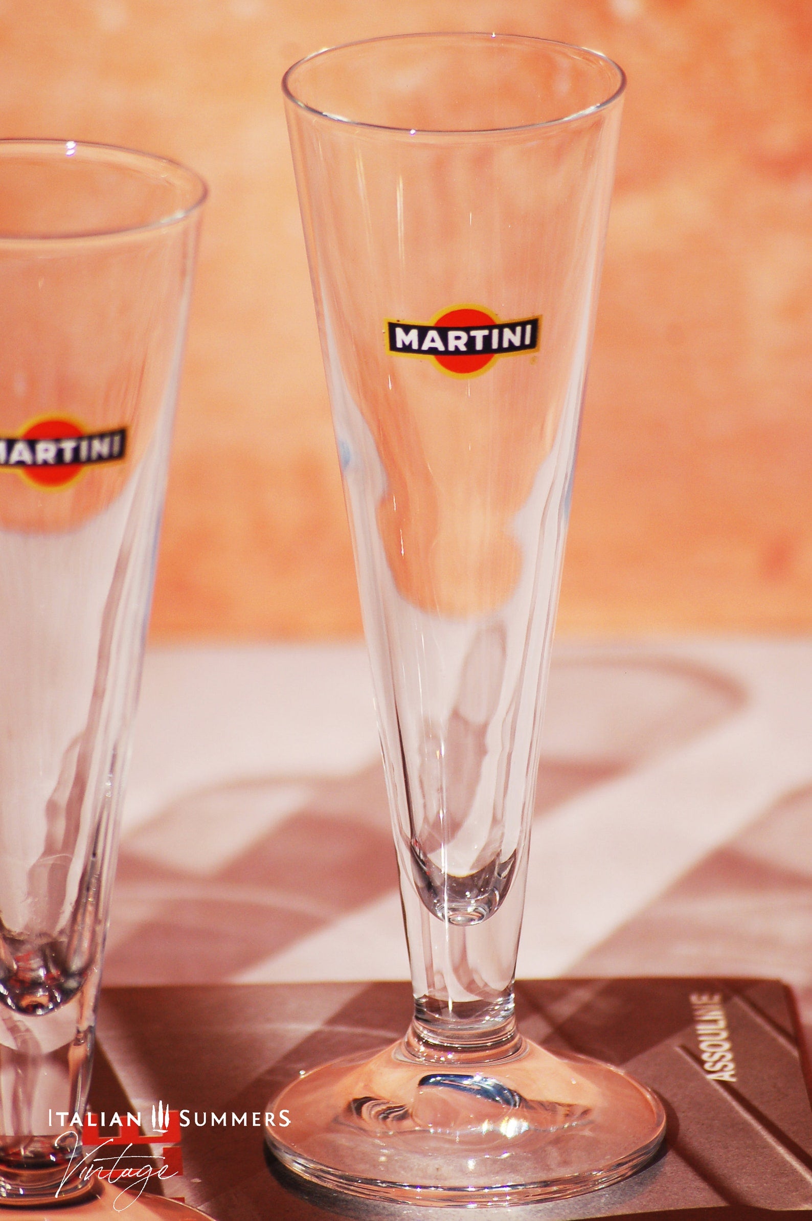 Set of four elegant vintage Martini glasses, each featuring the iconic Martini logo on the front. The glasses are displayed on a book titled 'Dolce Vita Style', set against a warm, peach-colored background with the text 'Italian Summers Vintage'.