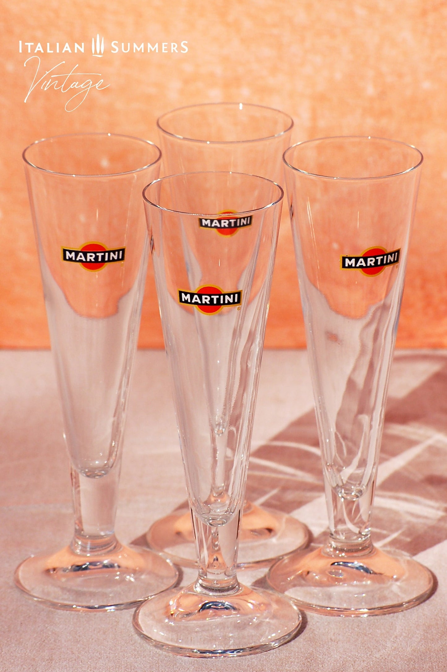 Set of four elegant vintage Martini glasses, each featuring the iconic Martini logo on the front. The glasses are displayed on a book titled 'Dolce Vita Style', set against a warm, peach-colored background with the text 'Italian Summers Vintage'.
