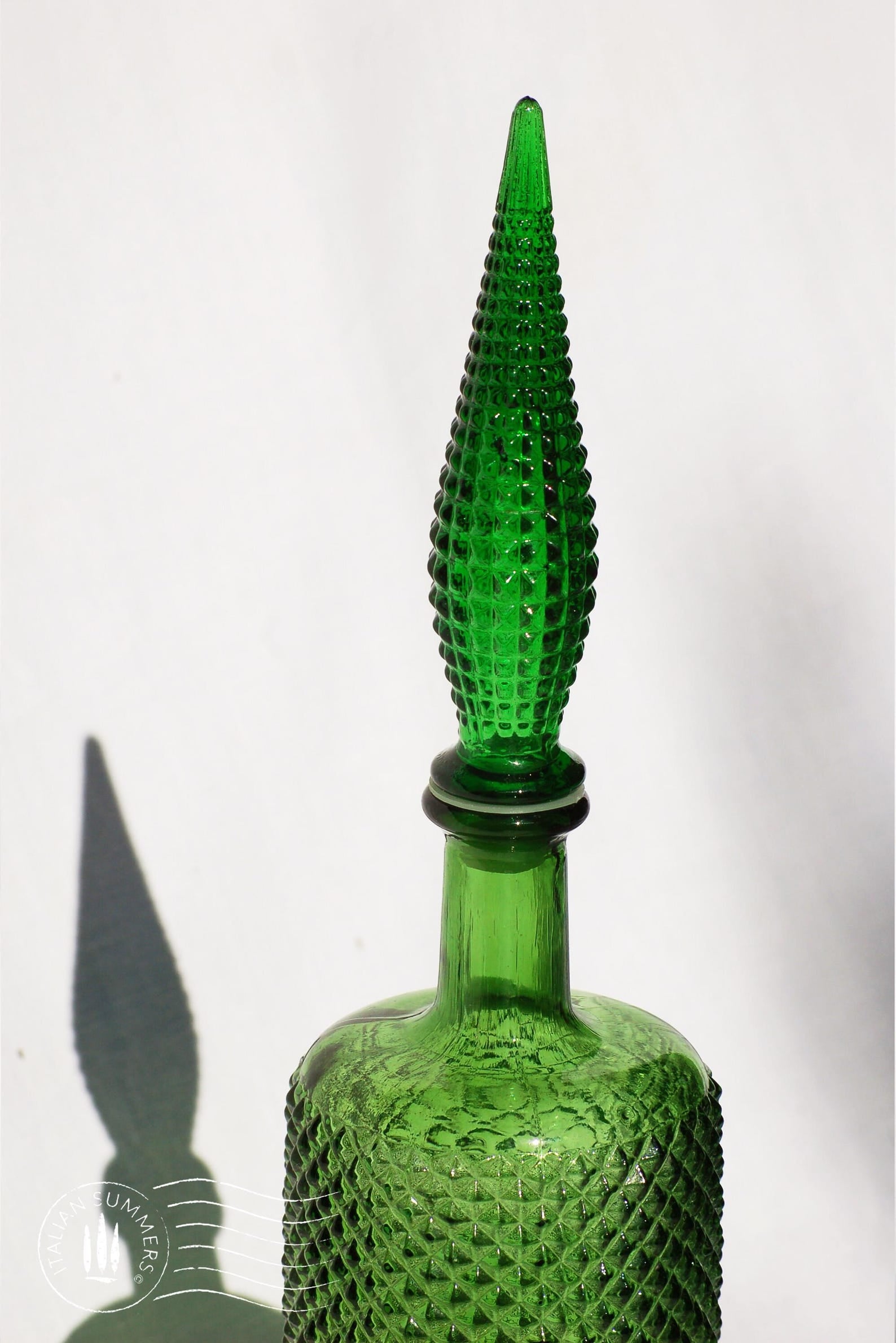 detail of a  large, tall green Empoli Glass Genie Bottle embossed with a diamonds pattern and a tall glass embossed flame-shaped stopper of the same material glistening in the sun against a white cloth background