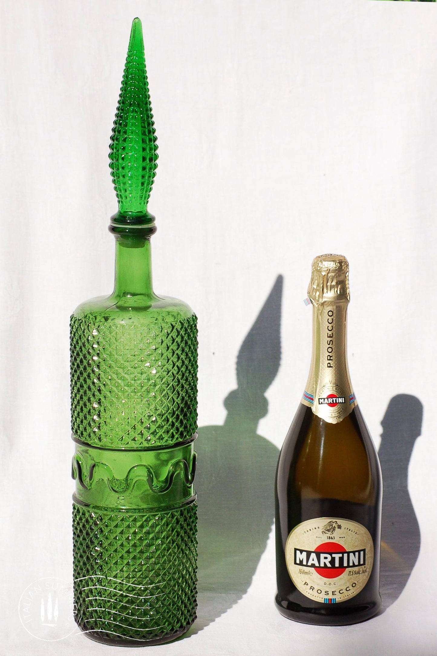 A large, tall green Empoli Glass Genie Bottle embossed with a diamonds pattern and a tall glass embossed flame-shaped stopper of the same material glistening in the sun against a white cloth background