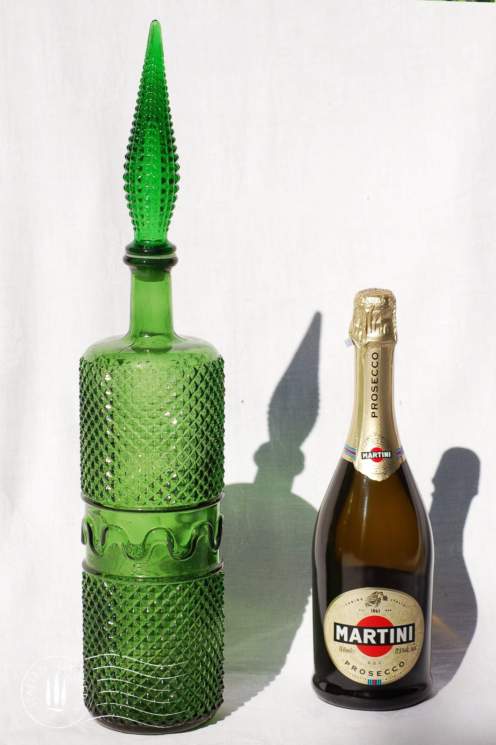 A large, tall green Empoli Glass Genie Bottle embossed with a diamonds pattern and a tall glass embossed flame-shaped stopper of the same material glistening in the sun against a white cloth background