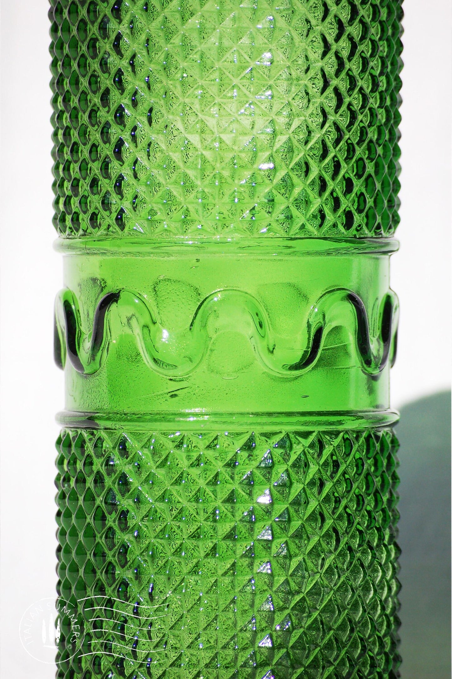 detail of a  large, tall green Empoli Glass Genie Bottle embossed with a diamonds pattern and a tall glass embossed flame-shaped stopper of the same material glistening in the sun against a white cloth background