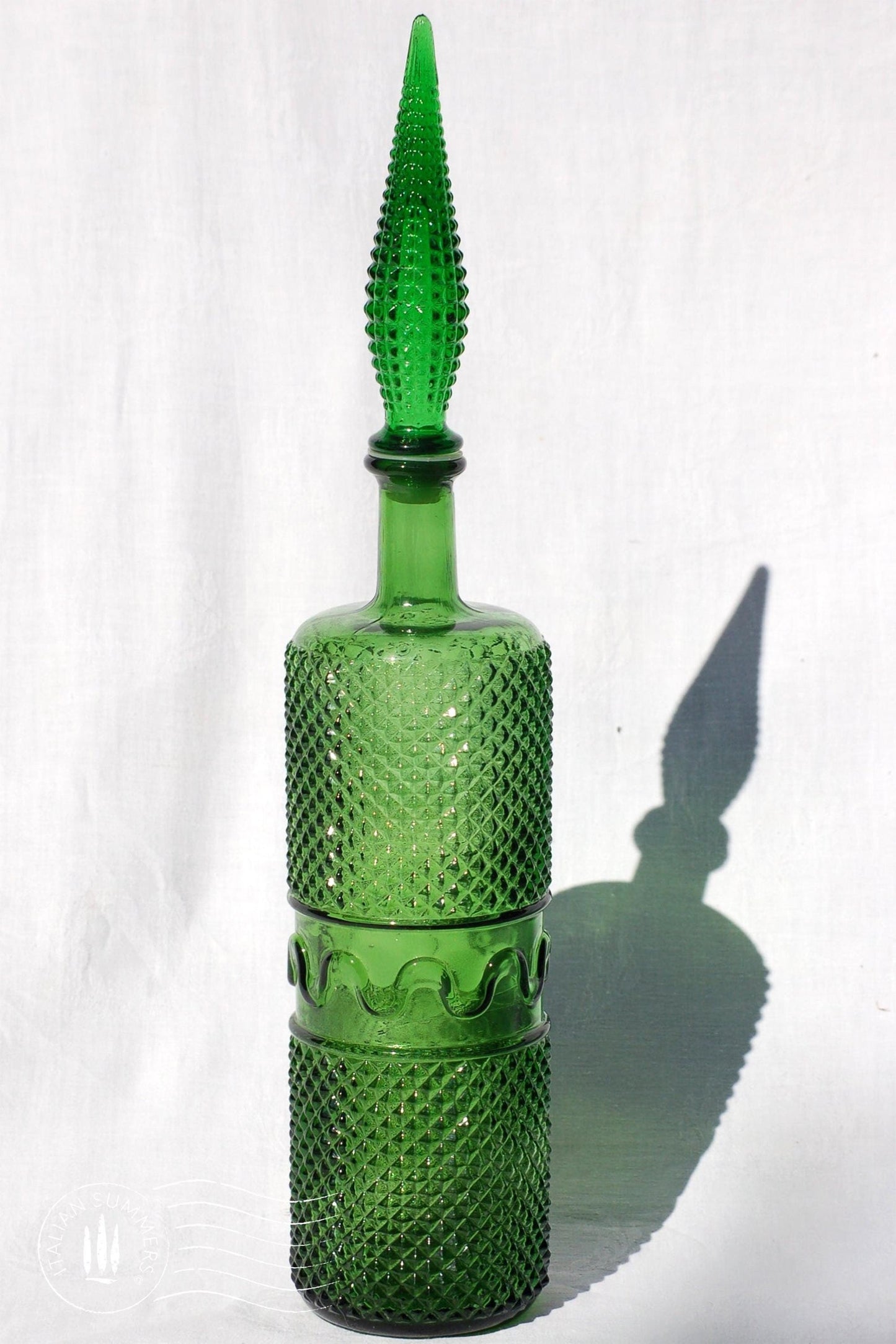A large, tall green Empoli Glass Genie Bottle embossed with a diamonds pattern and a tall glass embossed flame-shaped stopper of the same material glistening in the sun against a white cloth background