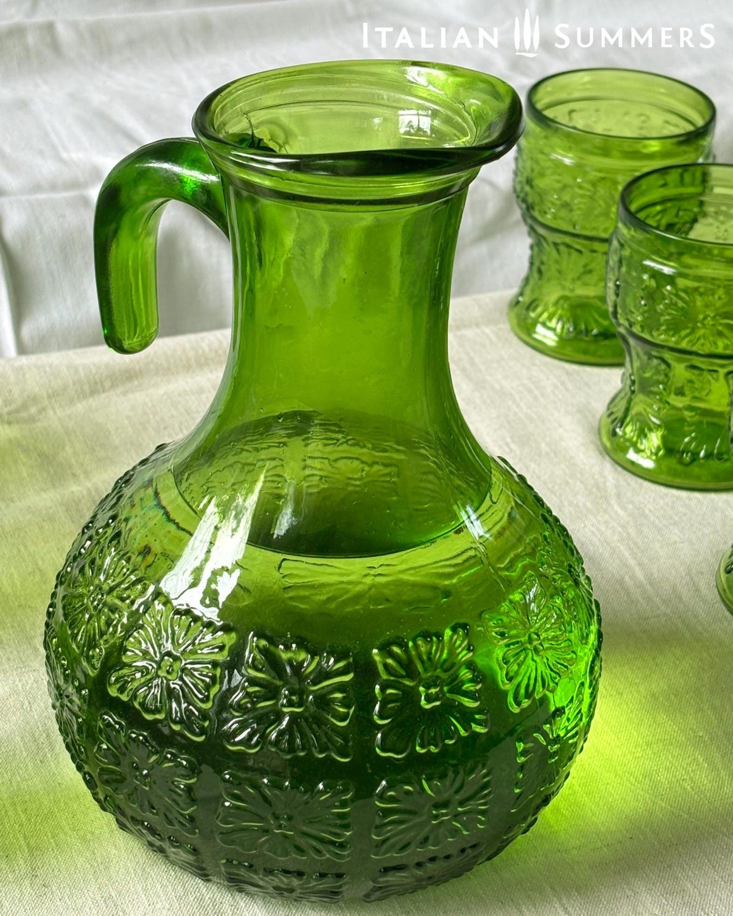 A green glass Italian carafe and six tumblers set on a white tablecloth