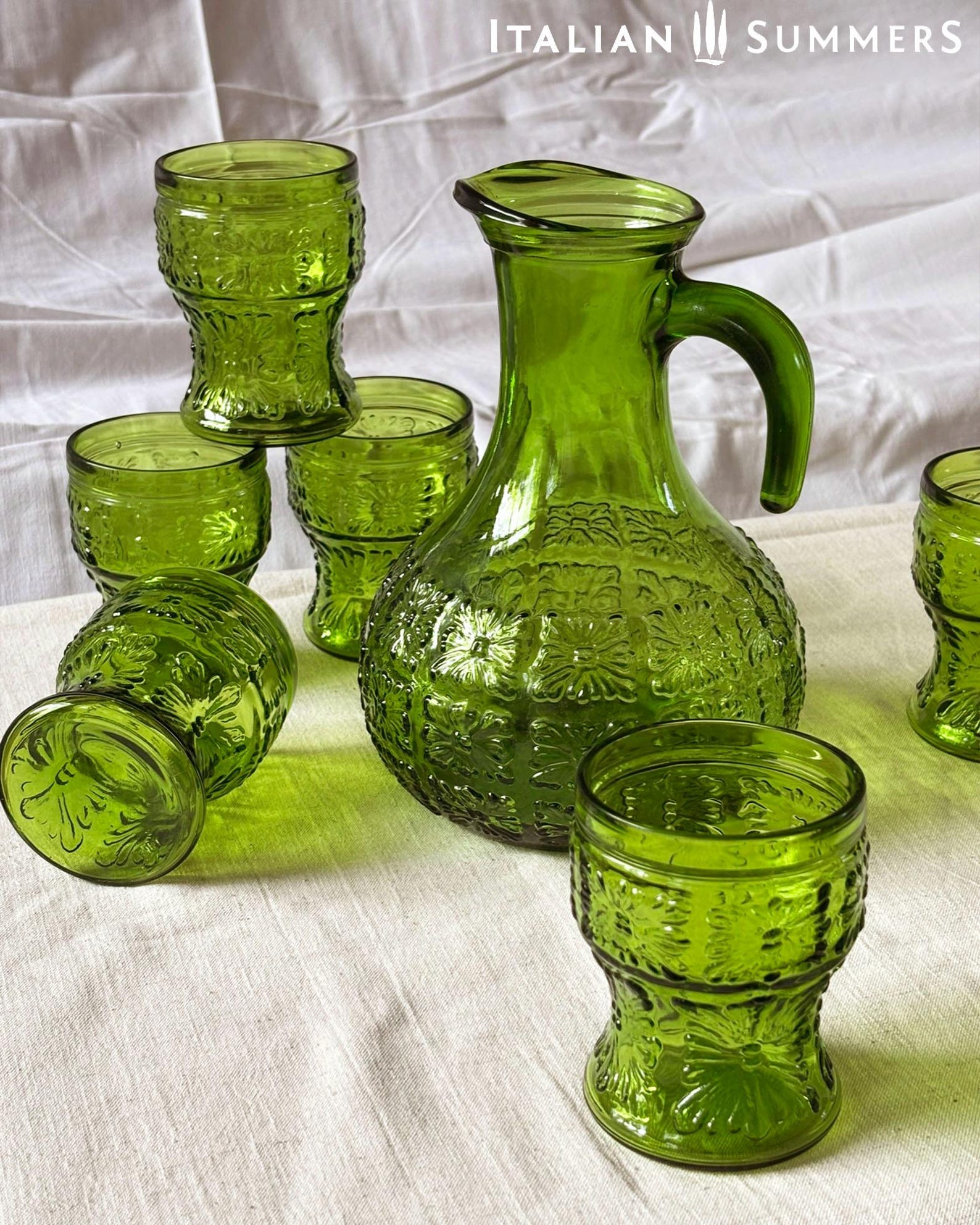 A green glass Italian carafe and six tumblers set on a white tablecloth