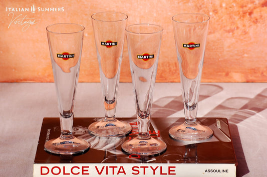 Set of four elegant vintage Martini glasses, each featuring the iconic Martini logo on the front. The glasses are displayed on a book titled 'Dolce Vita Style', set against a warm, peach-colored background with the text 'Italian Summers Vintage'.