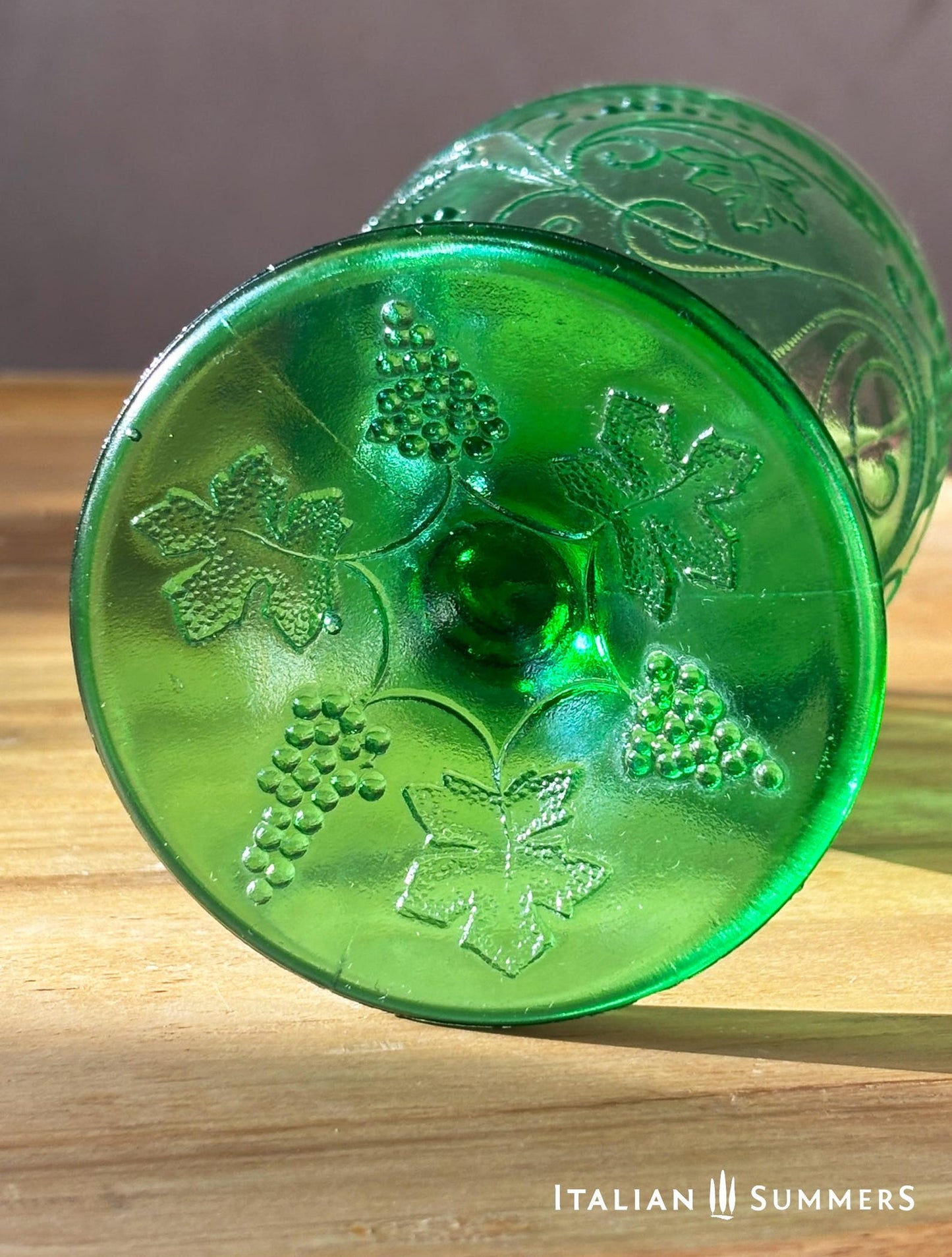 A rich emerald-green glass  set of six wine goblets with embossed grapes and vines motif from Tuscany on a red and white checkered tablecloth ad a terracotta wall in the background..