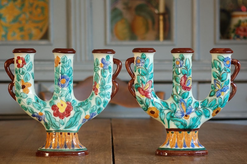 A pair of colorful ceramic candleholders by Cerart Monaco  side by side, both decorated with floral designs,  on a wooden surface