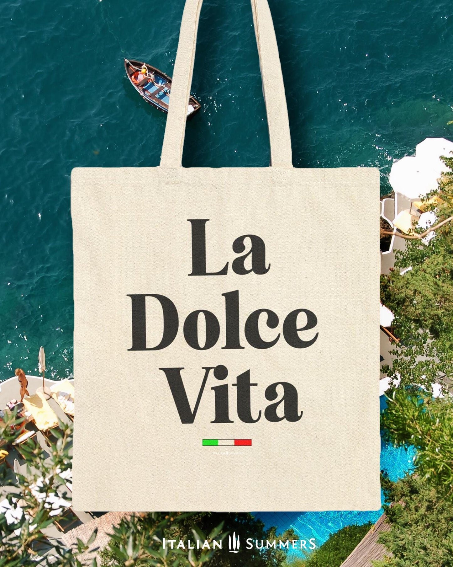 A cotton canvas tote bag with the text  La Dolce Vita  floats in front of an Idyllic Mediterranean seaside view.