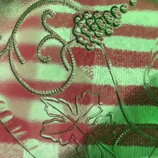 A rich emerald-green glass  set of six wine goblets with embossed grapes and vines motif from Tuscany on a red and white checkered tablecloth ad a terracotta wall in the background..