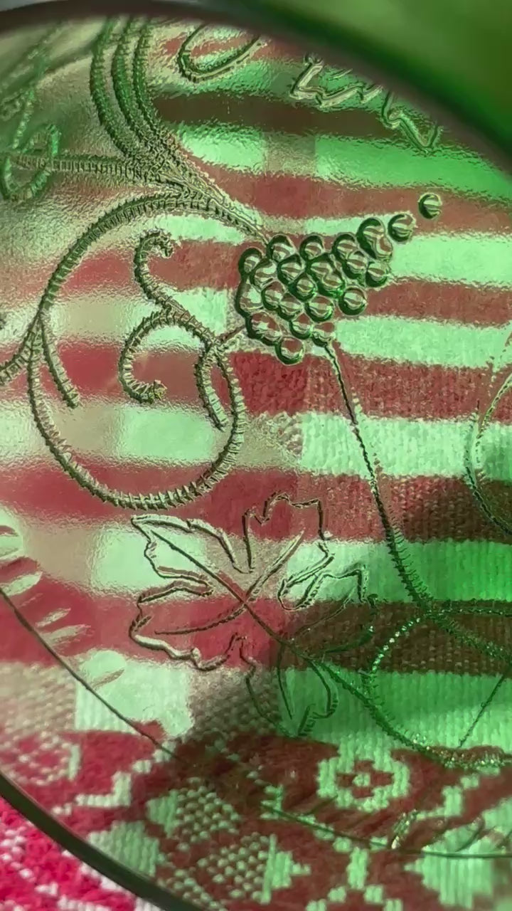 A rich emerald-green glass  set of six wine goblets with embossed grapes and vines motif from Tuscany on a red and white checkered tablecloth ad a terracotta wall in the background..