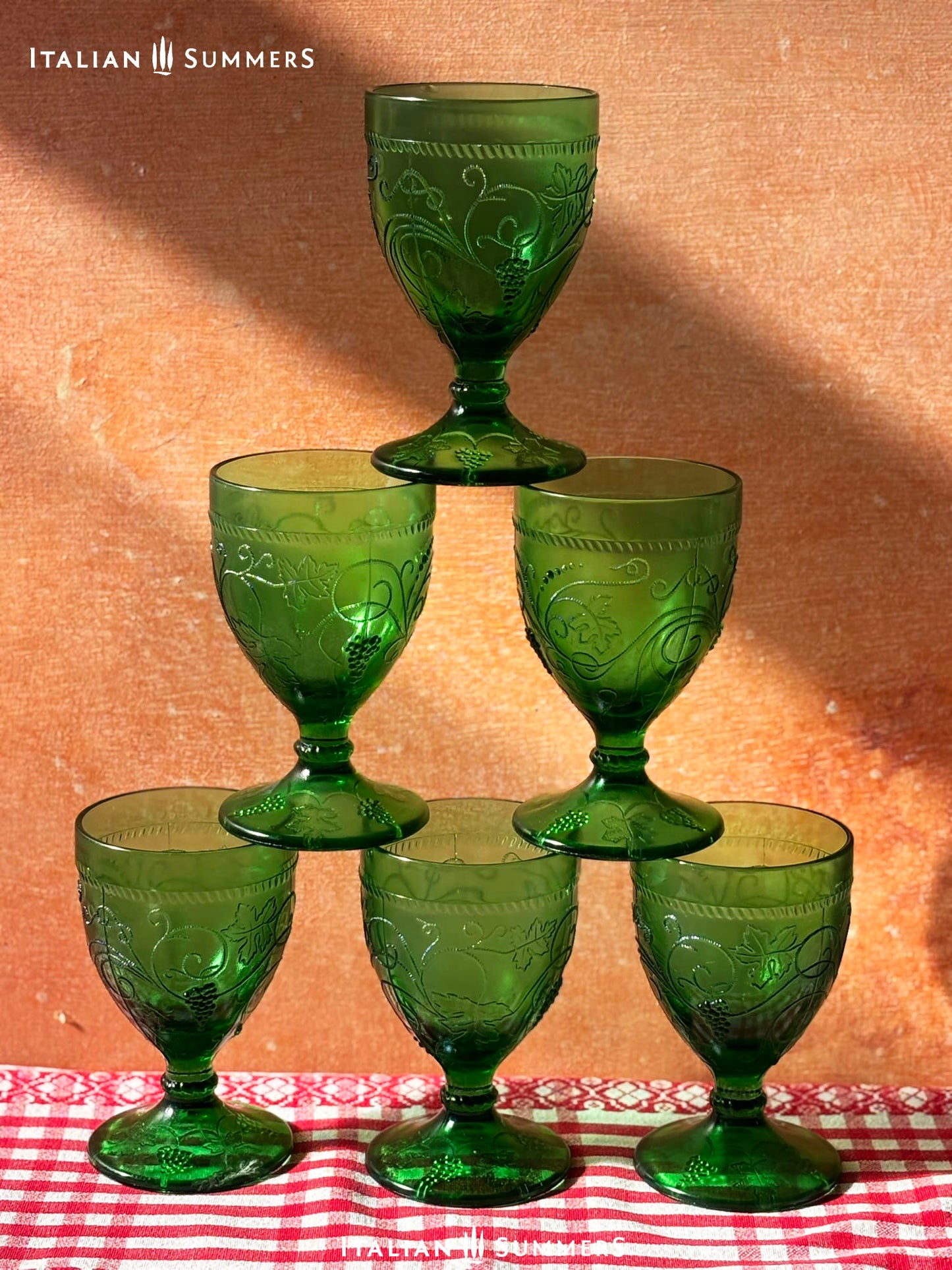 A rich emerald-green glass  set of six wine goblets with embossed grapes and vines motif from Tuscany on a red and white checkered tablecloth ad a terracotta wall in the background..