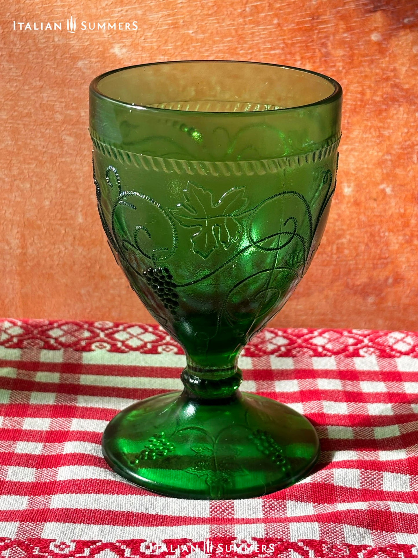Green glass goblet with intricate embossed grape and vines designs on a red and white checkered tablecloth, branded 'Italian Summers'.