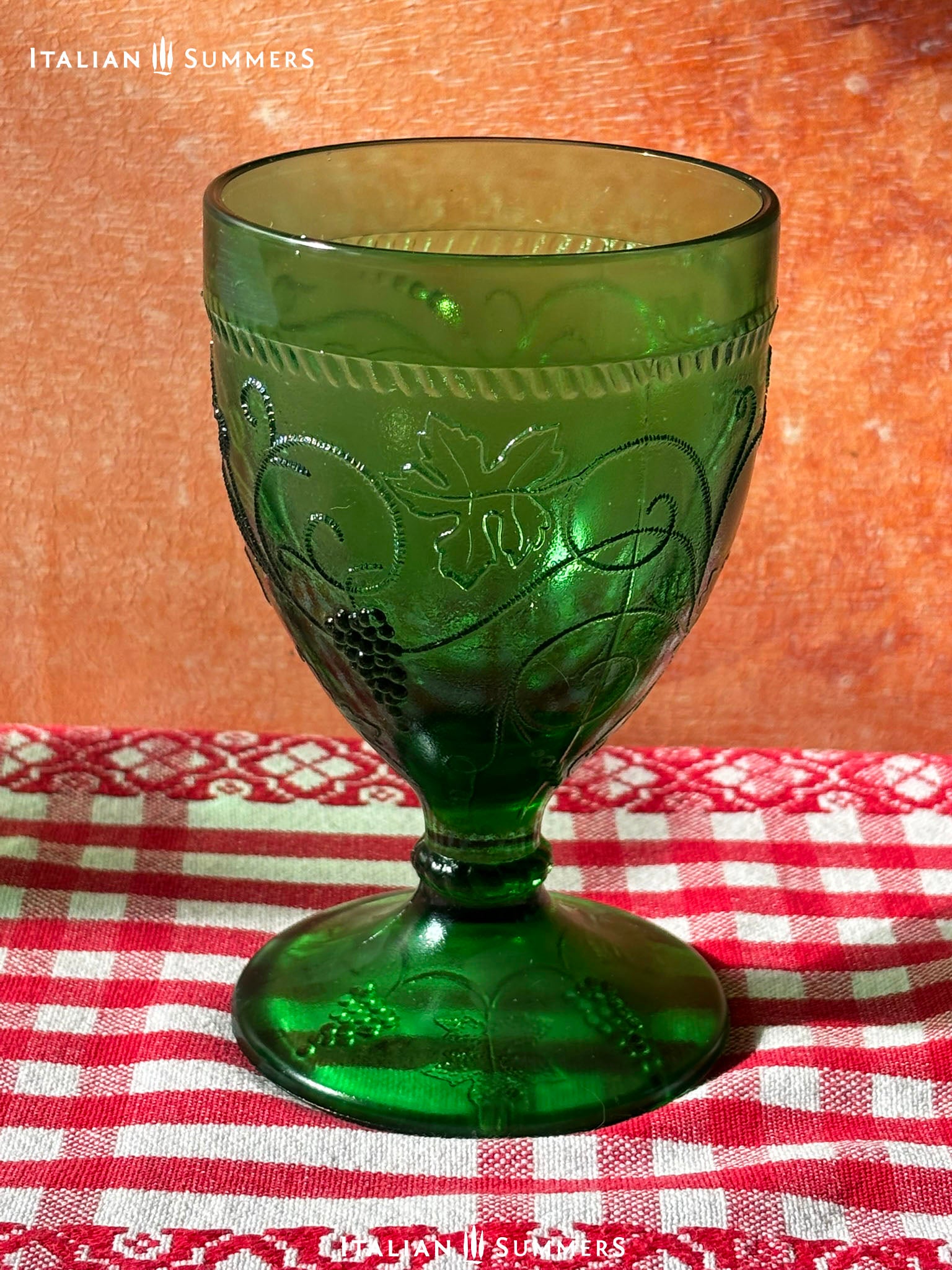 Green glass goblet with intricate embossed grape and vines designs on a red and white checkered tablecloth, branded 'Italian Summers'.
