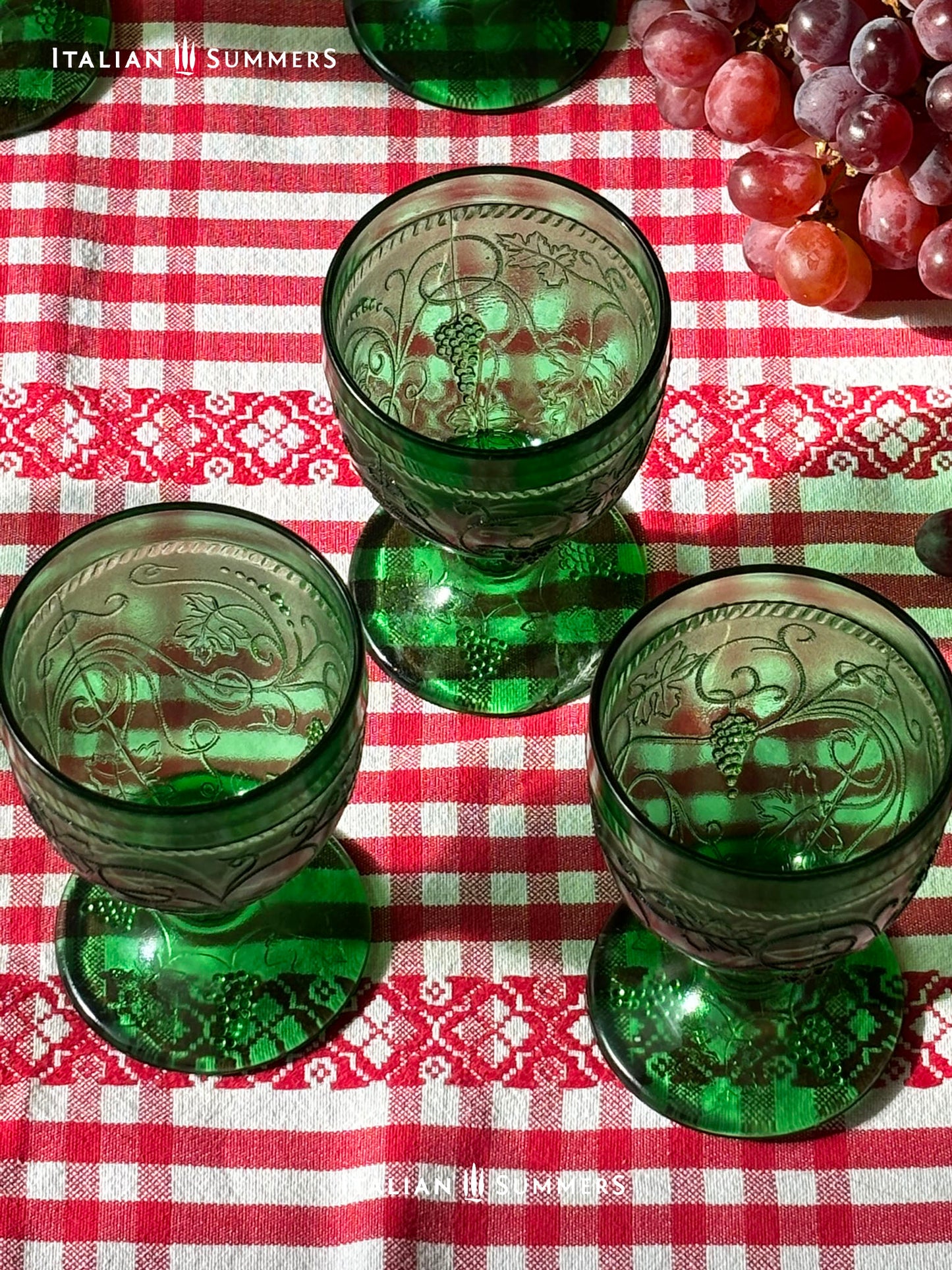 Green glass goblets with embossed designs on a red and white checkered tablecloth, accompanied by grapes.