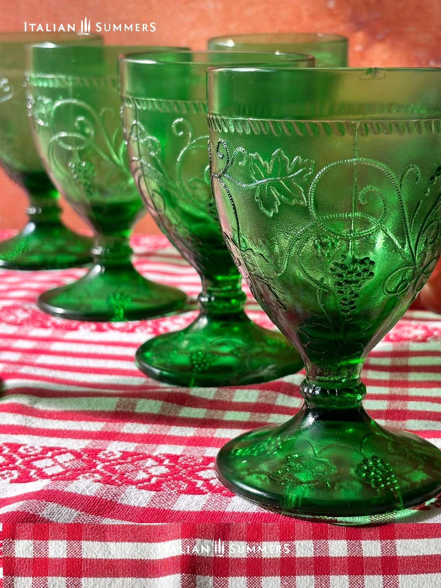 Green glass goblets with embossed grapes and vines designs on a red and white checkered tablecloth.