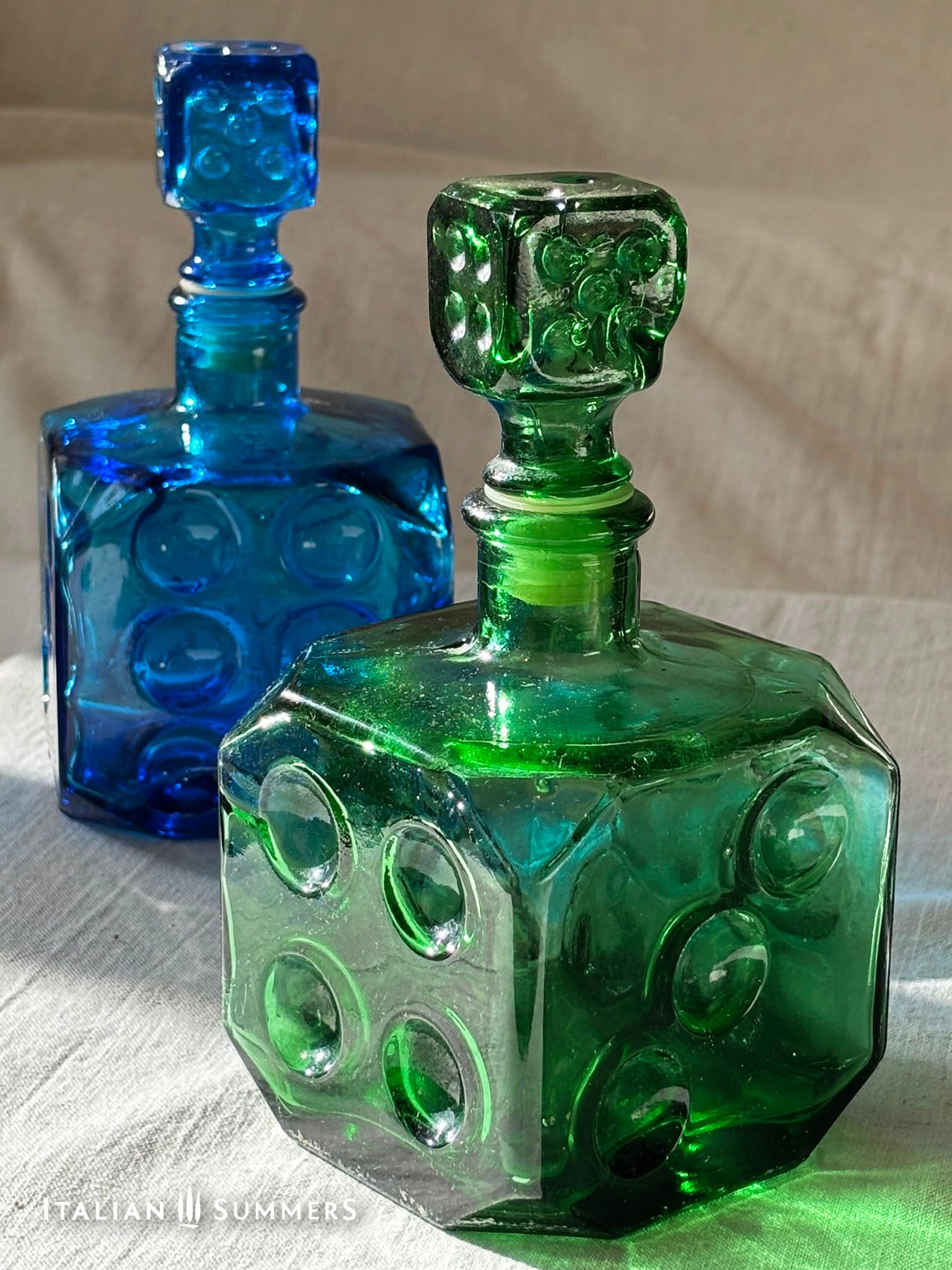 Two Emerald Green Empoli dice glass decanter on a light background, one blue and one green, with decorative stoppers
