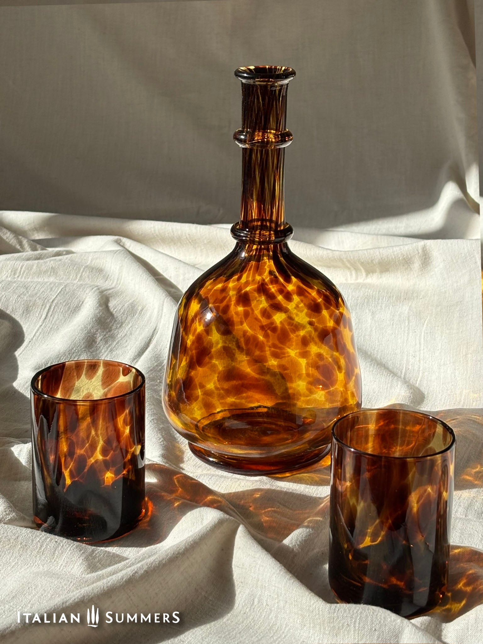 Tortoiseshell glassware including a bottle, two glasses on a white fabric background.