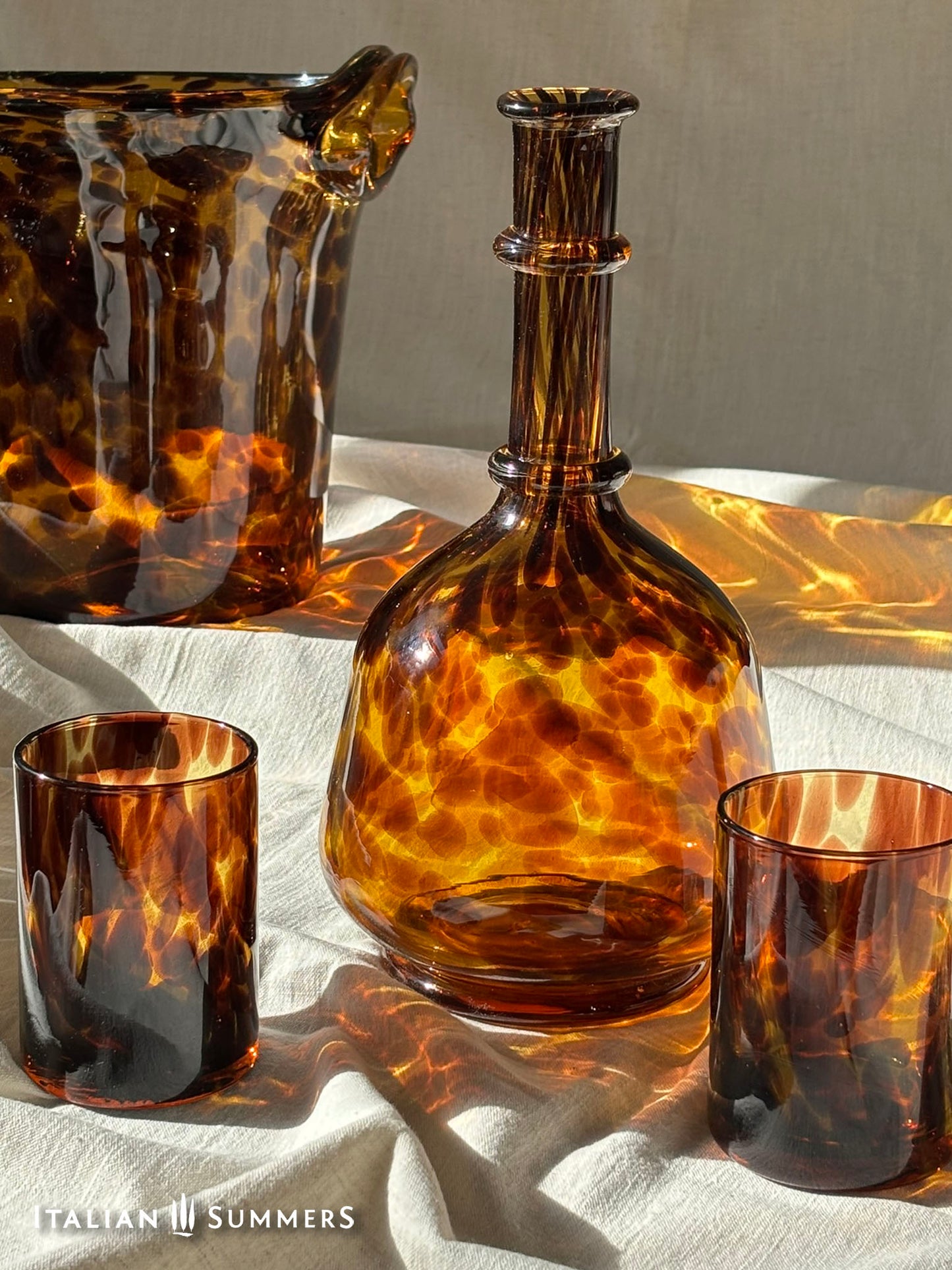 Tortoiseshell glassware including a bottle, two glasses, and an ice bucket on a white fabric background.