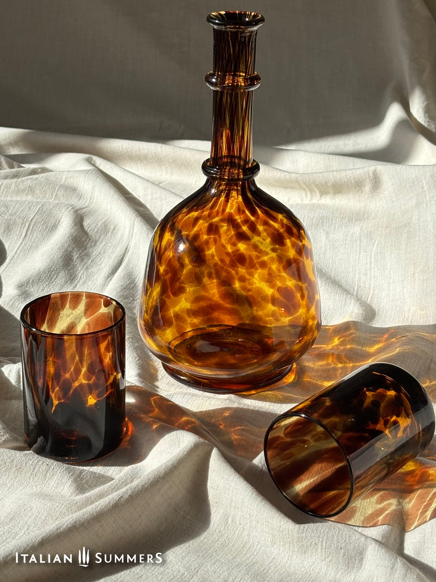 Tortoiseshell-patterned glass decanter, glass, and carafe on a white fabric background with 'Italian Summers' branding.