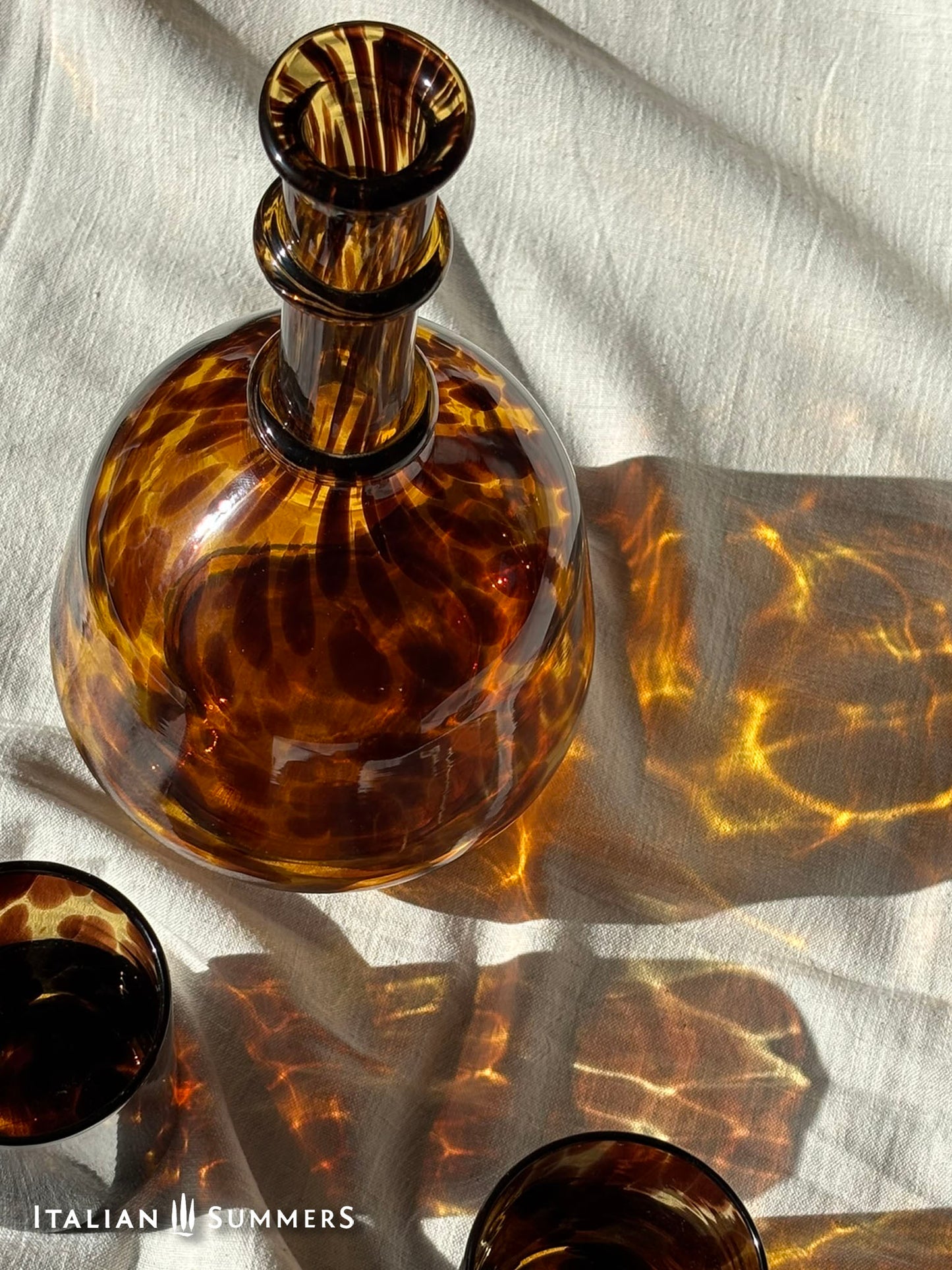 Tortoiseshell-patterned glass decanter on a textured fabric background with 'Italian Summers' branding.
