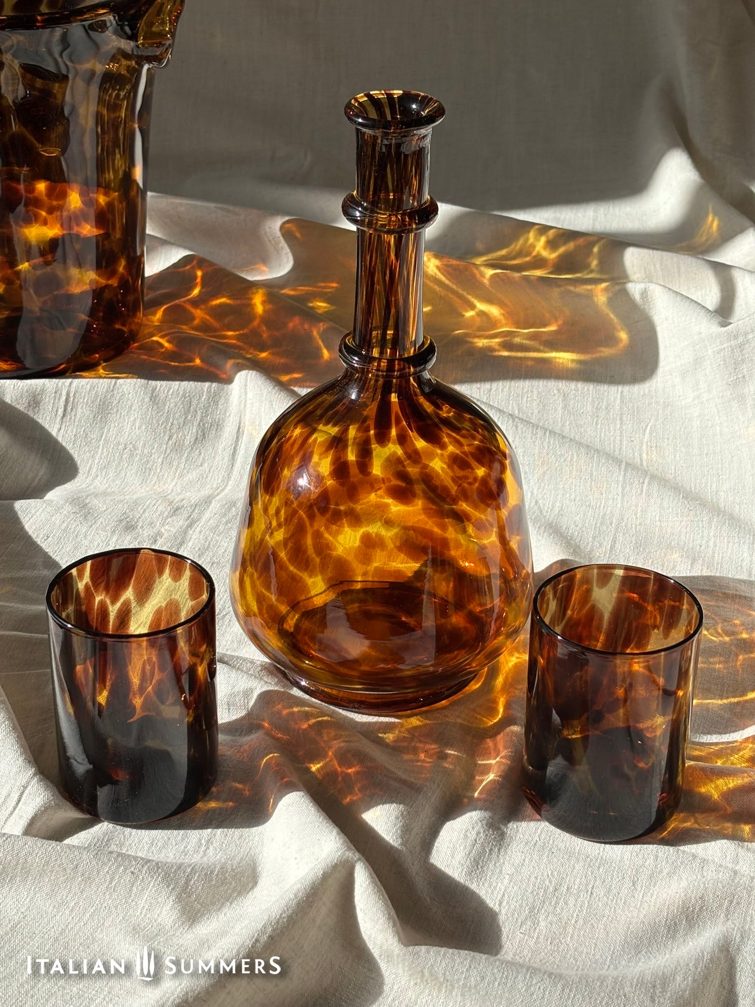 Tortoiseshell glassware including a bottle, two glasses on a white fabric background.