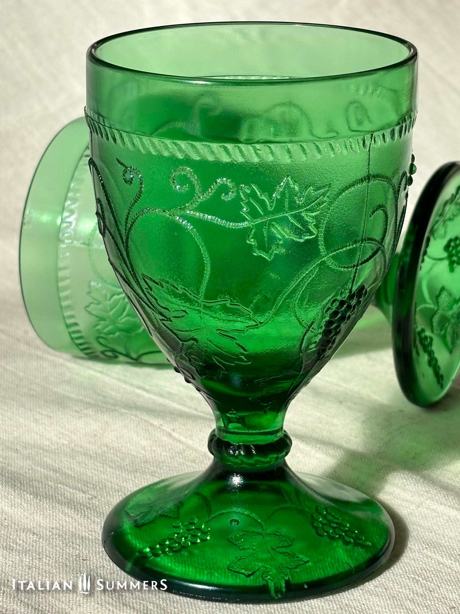 A rich emerald-green glass  set of six wine goblets with embossed grapes and vines motif from Tuscany on a red and white checkered tablecloth ad a terracotta wall in the background..
