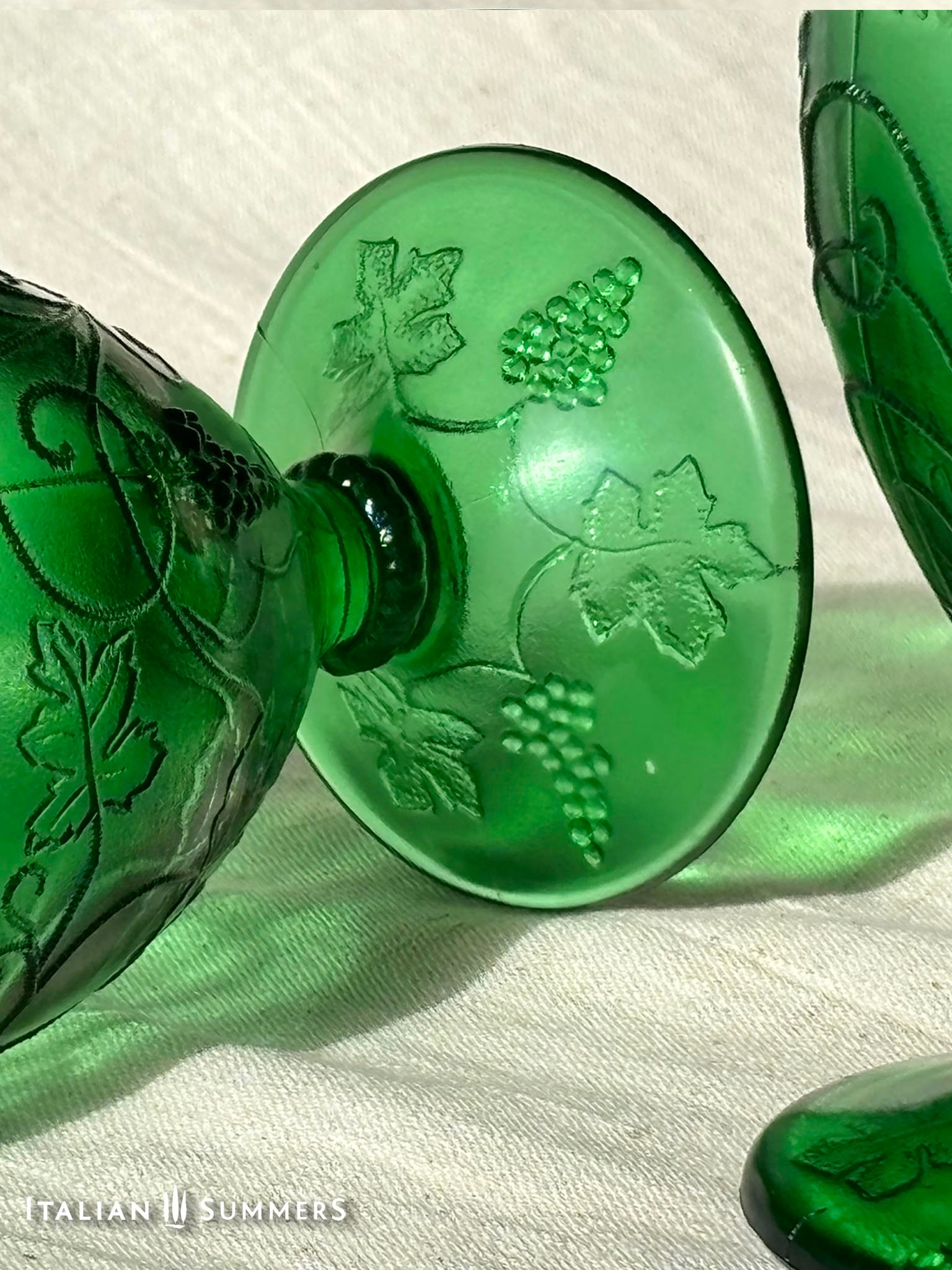 A rich emerald-green glass  set of six wine goblets with embossed grapes and vines motif from Tuscany on a red and white checkered tablecloth ad a terracotta wall in the background..